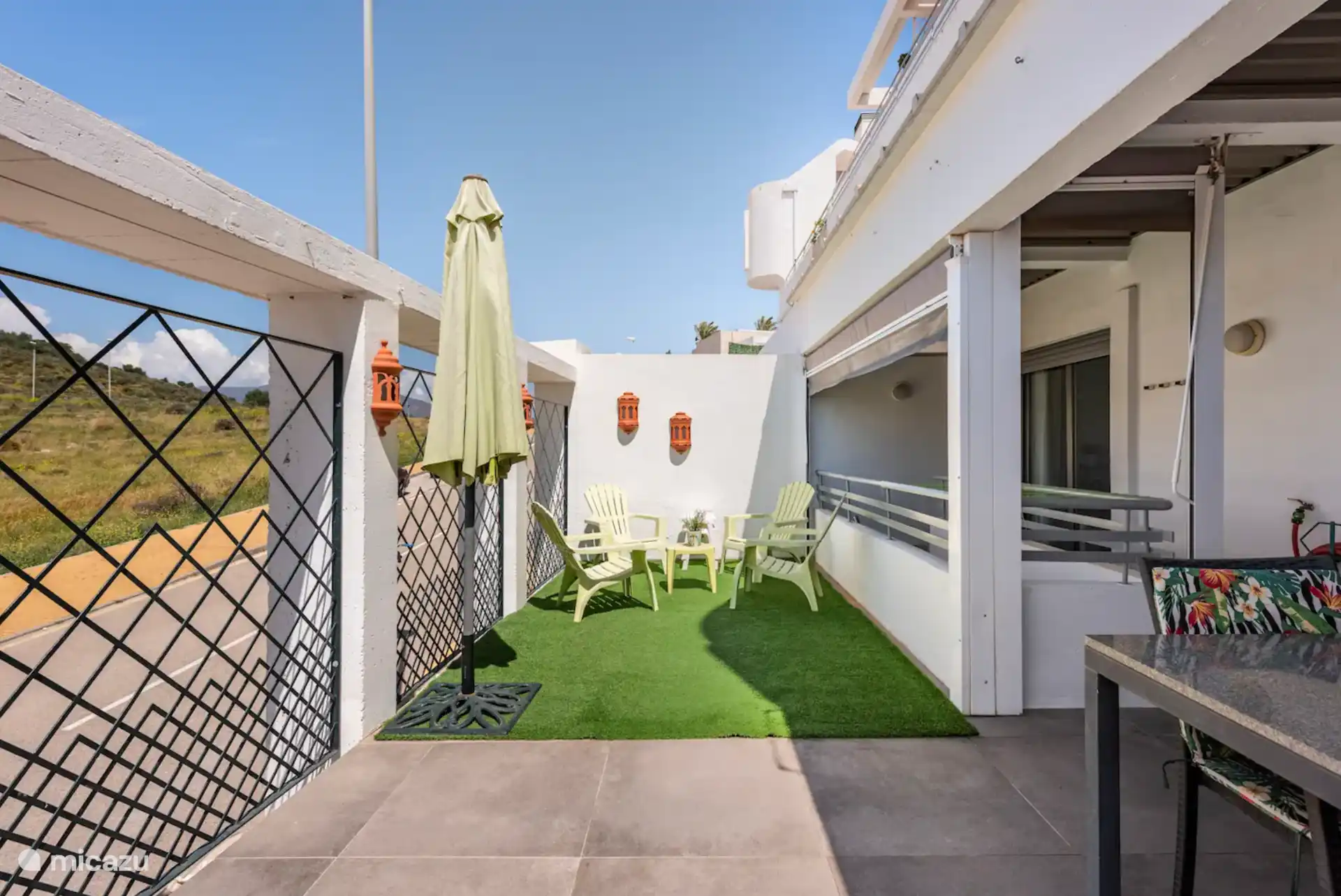 Terrasse sud-ouest prise sous un angle différent avec table pour 6 personnes, coin salon partiellement couvert sur gazon artificiel. Accès par le salon, la chambre principale et les escaliers à la troisième chambre et au parking privé. 