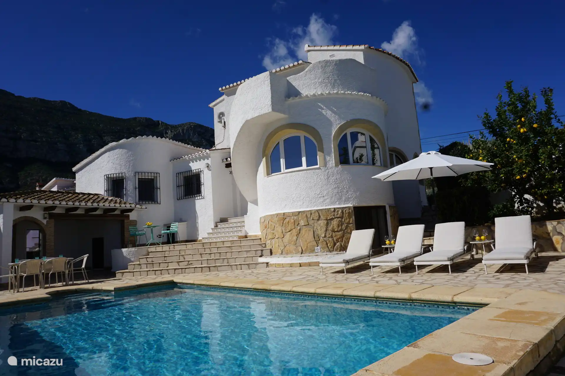 Parte trasera de nuestra casa con muchos asientos, rodeada de un jardín tropical con palmeras y árboles frutales. Los limones de tu propio jardín hacen que tu agua fría sepa muy bien.