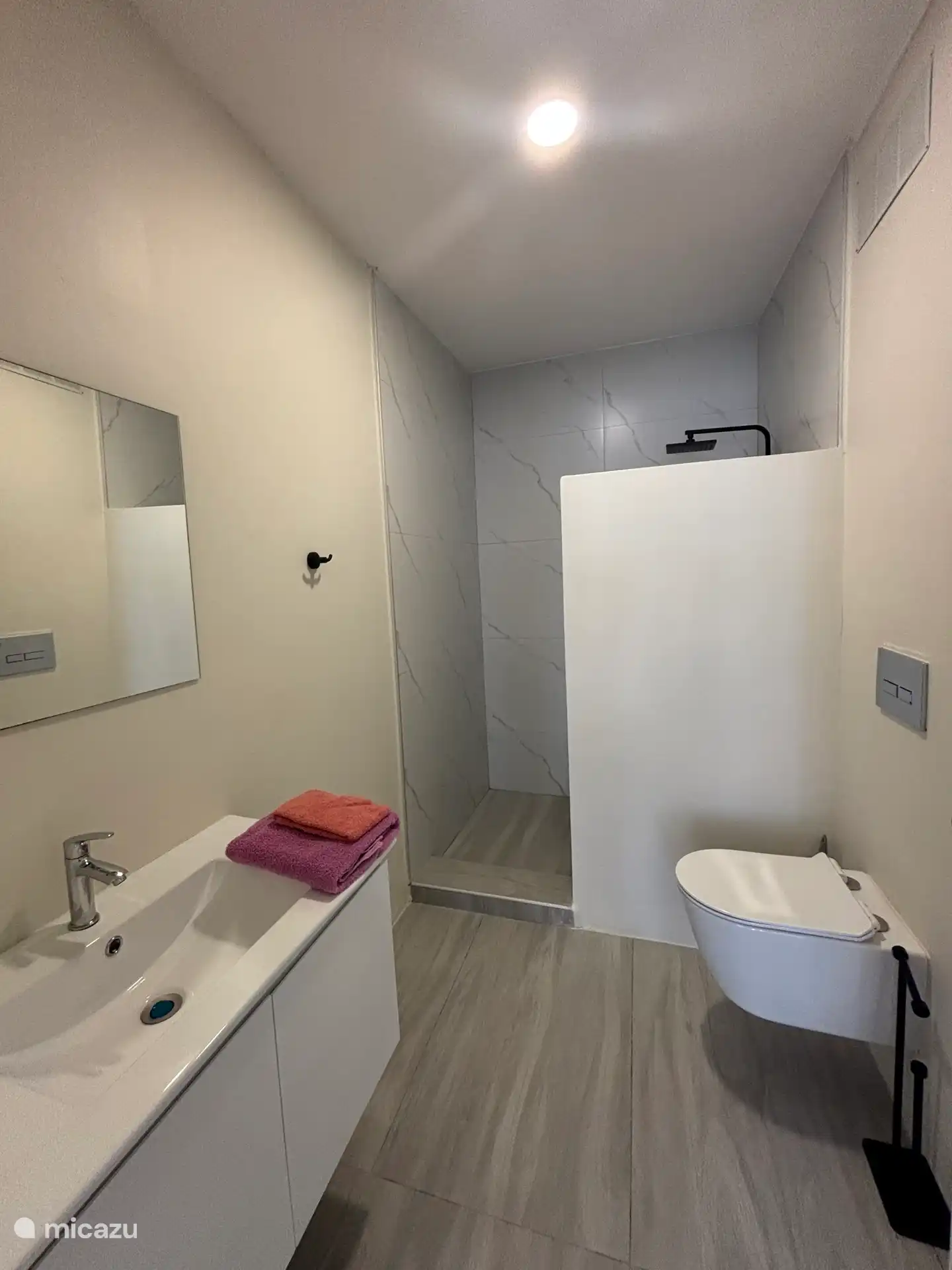 Geräumiges Badezimmer mit hängendem WC und einer ebenerdigen Dusche mit Regendusche. Ein Waschbecken mit einem Schrank. Steckdose und Haartrockner vorhanden. 