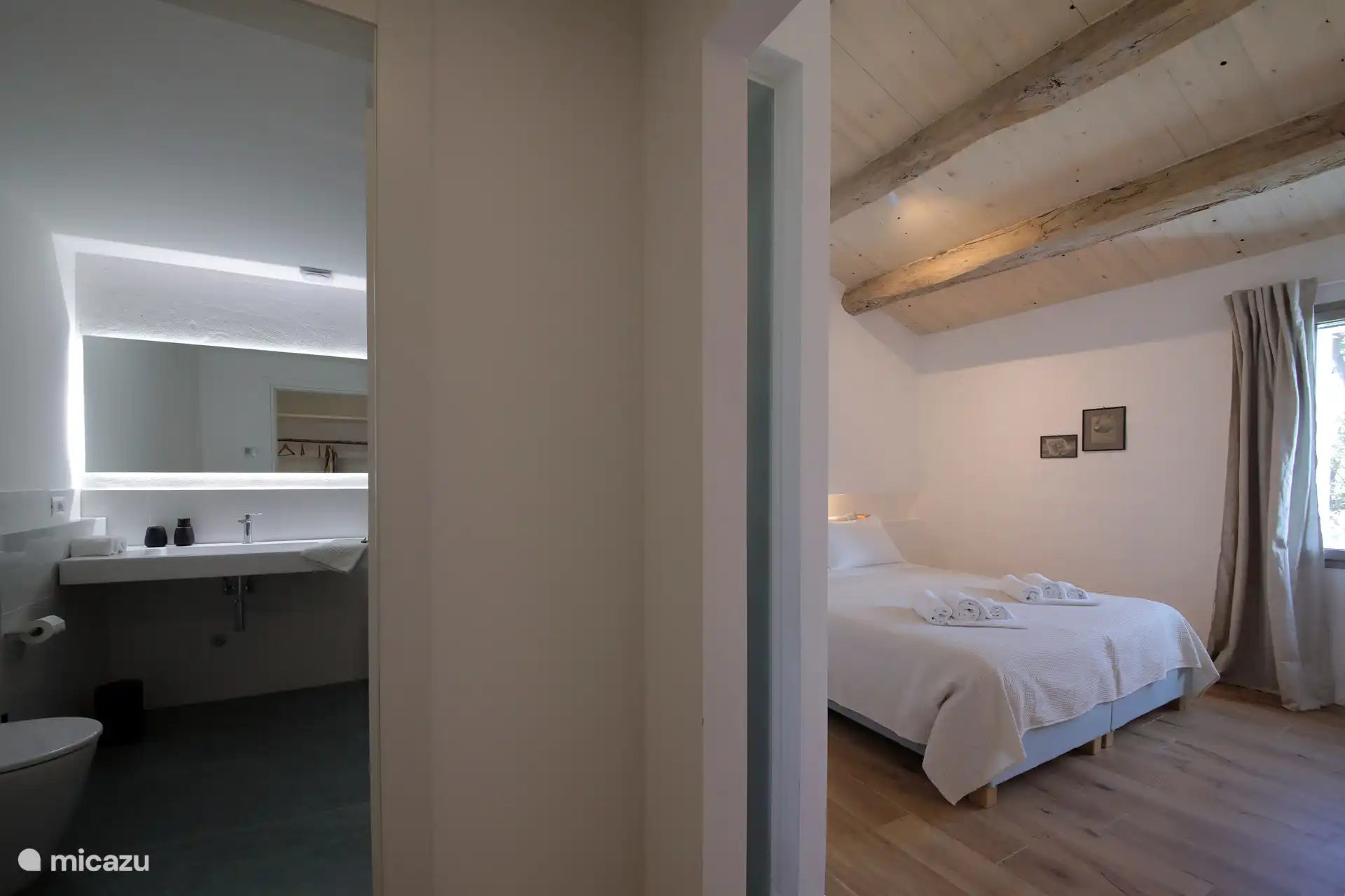 Ein helles, minimalistisches Schlafzimmer mit rustikal-schickem Flair und einer hohen geneigten Decke mit freiliegenden Holzbalken. Leichte Holzböden und weiße Wände schaffen eine luftige und entspannte Atmosphäre.