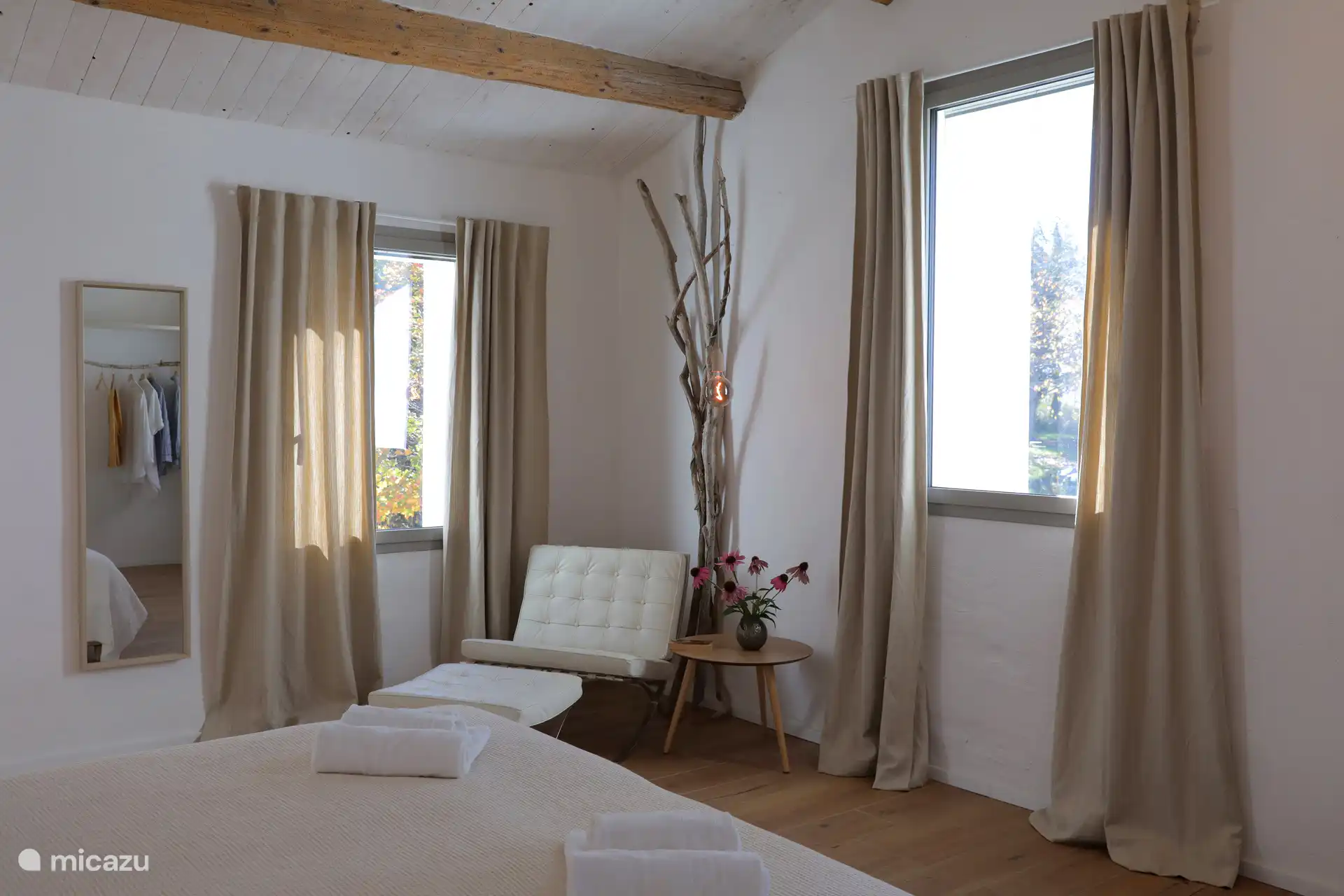 Ein helles, minimalistisches Schlafzimmer mit rustikal-schickem Flair und einer hohen geneigten Decke mit freiliegenden Holzbalken. Leichte Holzböden und weiße Wände schaffen eine luftige und entspannte Atmosphäre.