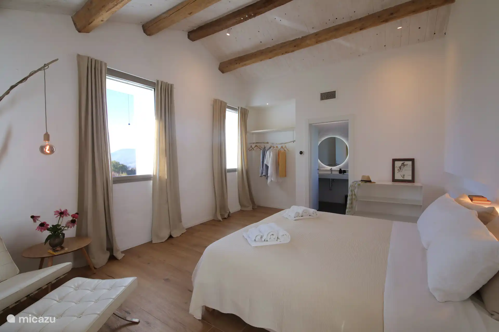 Ein helles, minimalistisches Schlafzimmer mit rustikal-schickem Flair und einer hohen geneigten Decke mit freiliegenden Holzbalken. Leichte Holzböden und weiße Wände schaffen eine luftige und entspannte Atmosphäre.