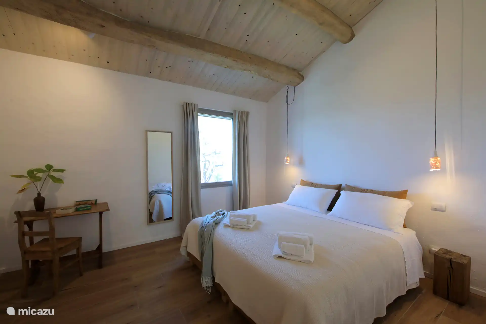 Ein helles, minimalistisches Schlafzimmer mit rustikal-schickem Flair und einer hohen geneigten Decke mit freiliegenden Holzbalken. Leichte Holzböden und weiße Wände schaffen eine luftige und entspannte Atmosphäre.