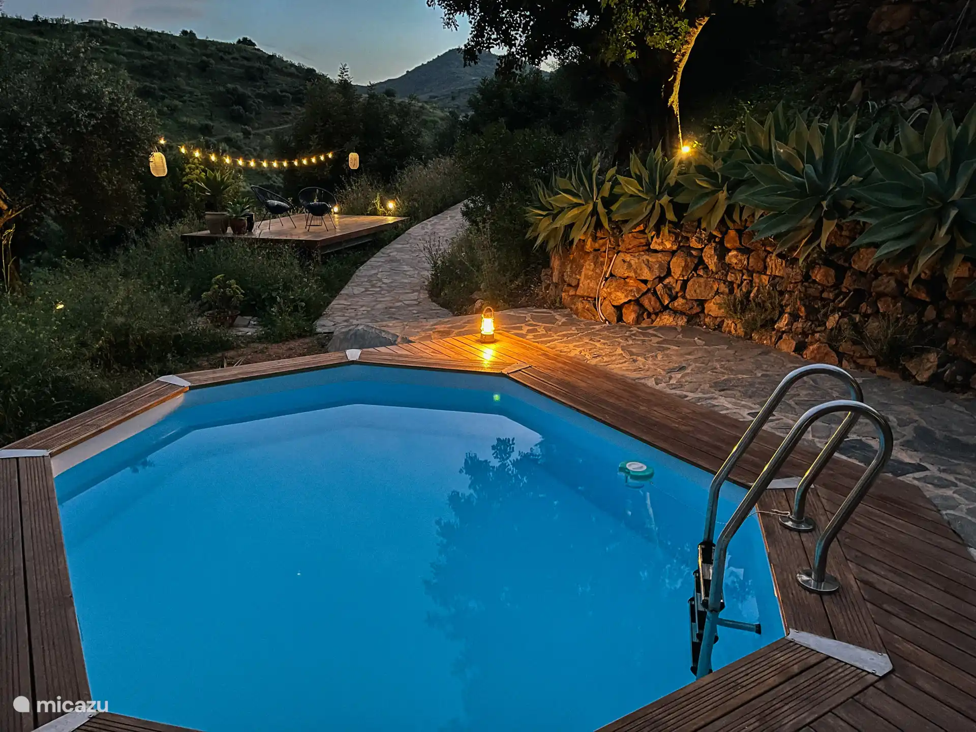 piscine la nuit avec derrière la terrasse en bois avec 2 fauteuils paresseux, une table et un éclairage d’ambiance