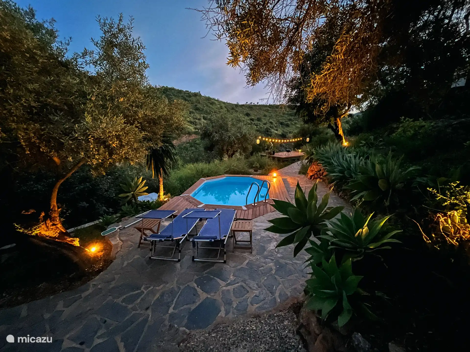Piscine la nuit avec des fauteuils longs, derrière elle la terrasse en bois avec 2 fauteuils paresseux, une table et un éclairage d’ambiance