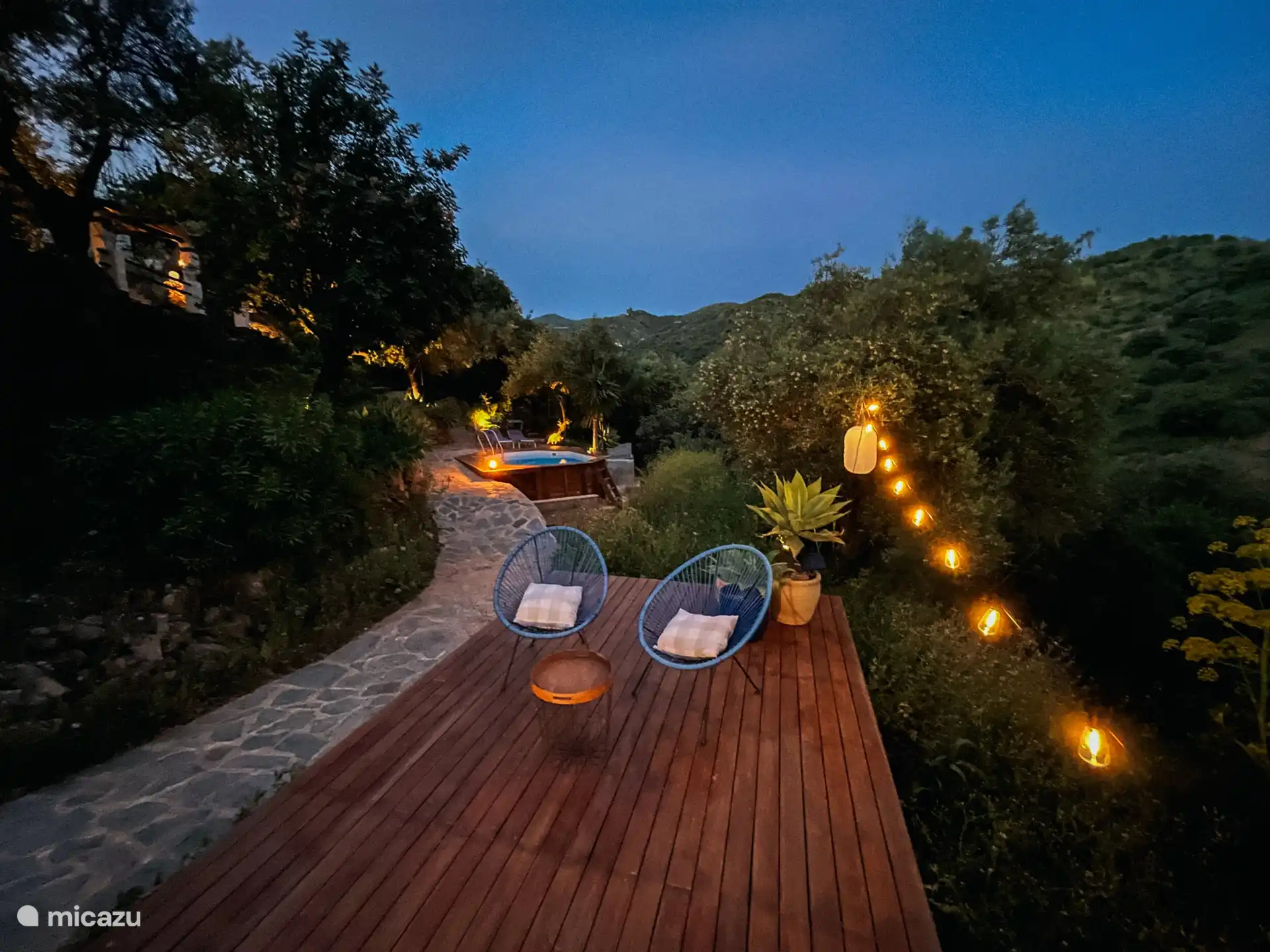 Terrasse en bois avec 2 fauteuils paresseux, table et éclairage d’ambiance, derrière elle la piscine 