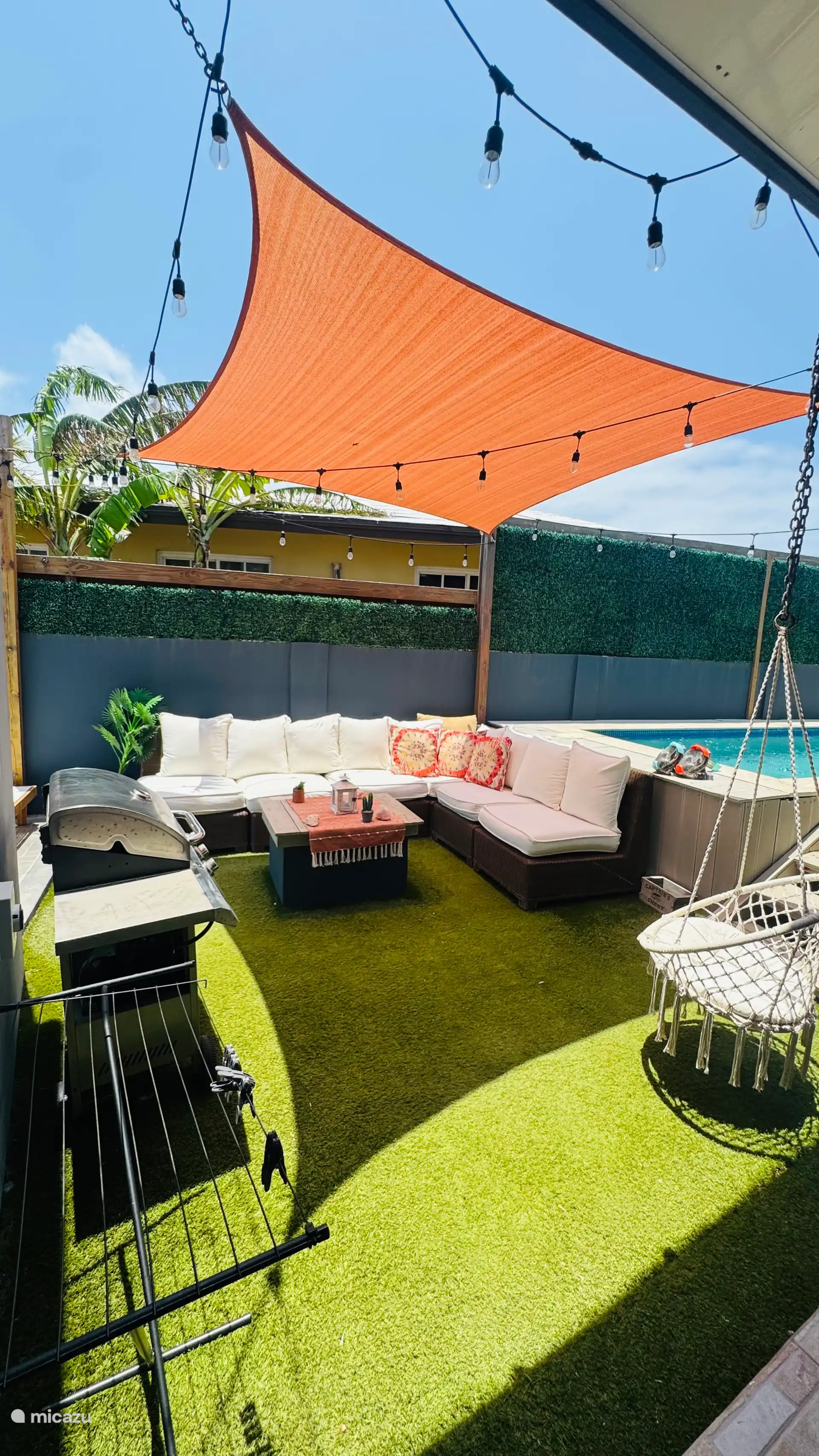 In het midden staat een comfortabele L-vormige buitenbank met lichtgekleurde kussens en een paar accentkussens met patroon, opgesteld rond een kleine houten salontafel.
BBQ