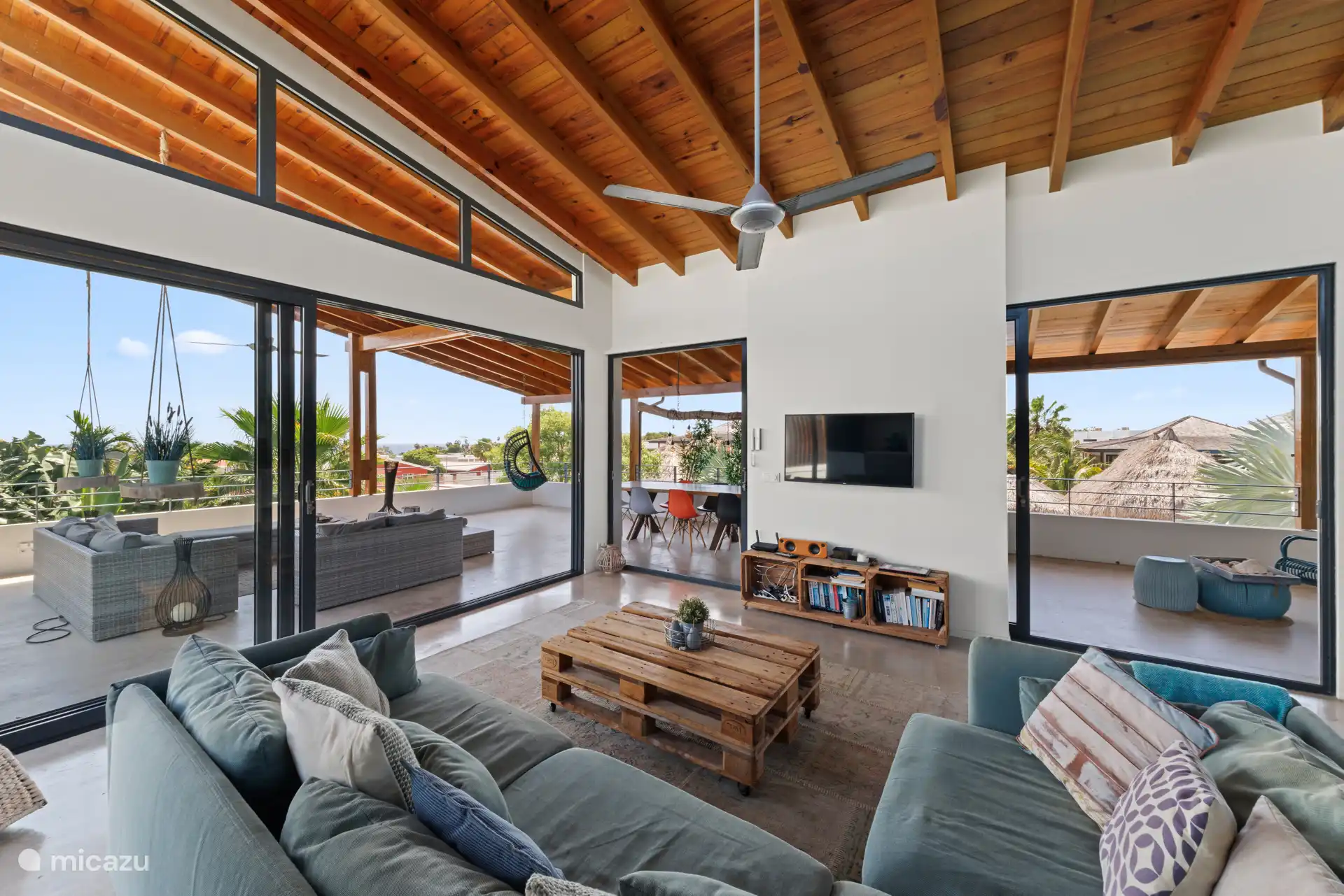 Woonkamer met hoog houten plafond, grote bank en TV. Veel licht door de grote ramen en stalen schuifdeuren 