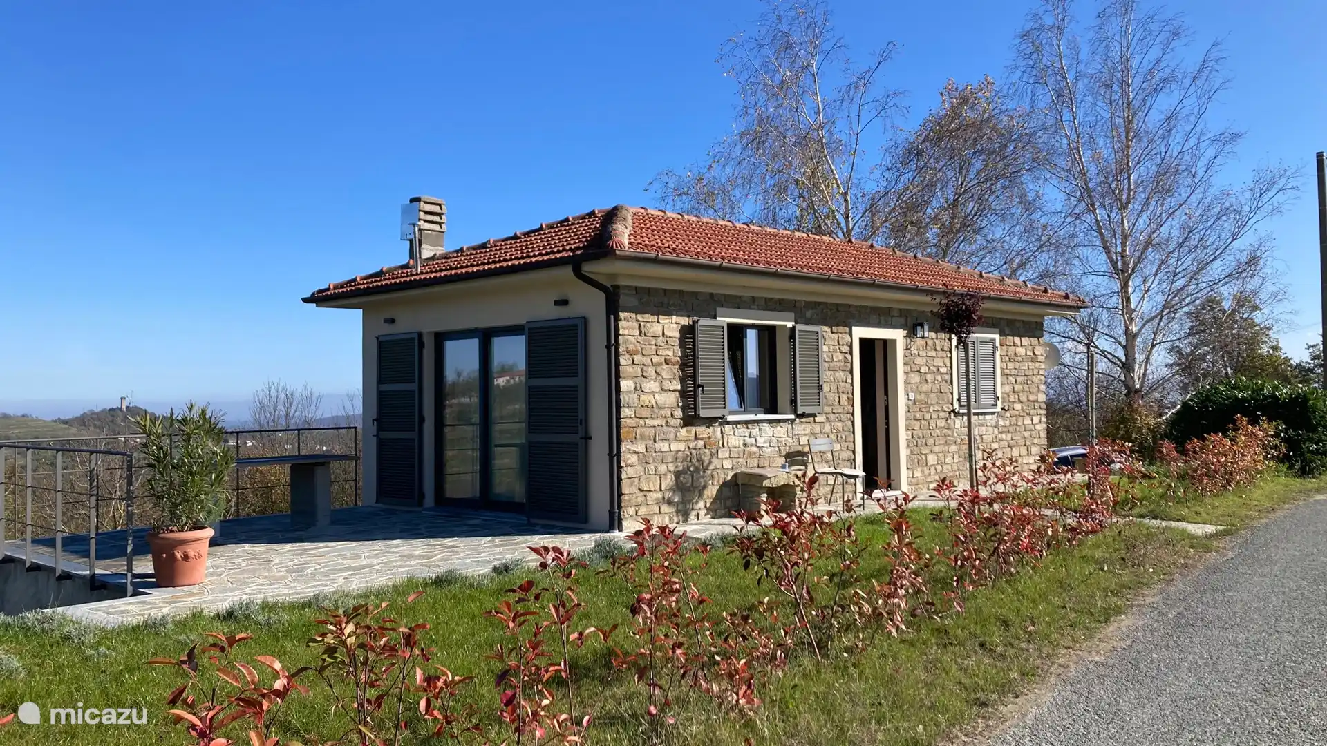 La maison à l’avant. Par les portes coulissantes, on entre dans la cuisine / le salon. Il y a 4 fauteuils de table à manger sur cette terrasse extérieure.