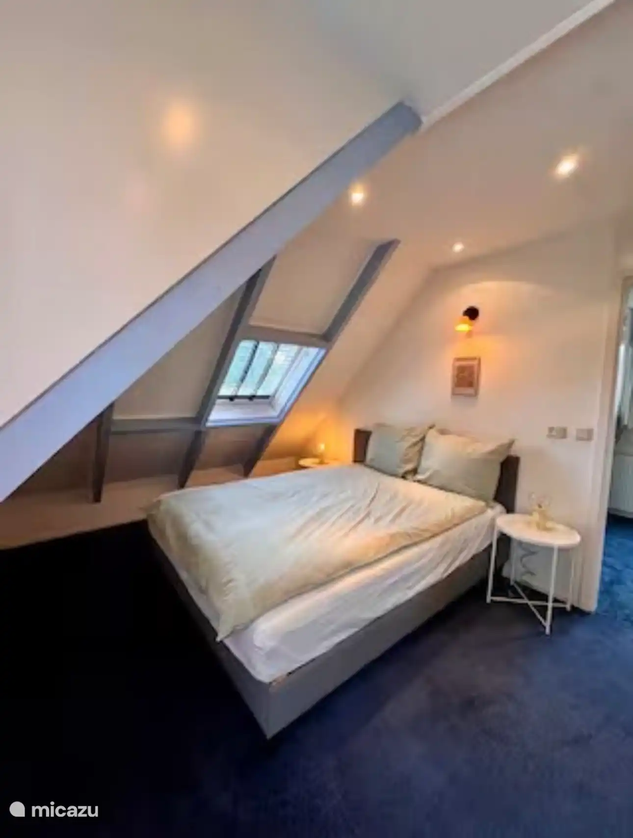 Knusse slaapkamer onder het schuine dak met warm licht en een dakraam. De zachte kleuren en rustige sfeer maken dit een fijne plek om helemaal tot rust te komen.