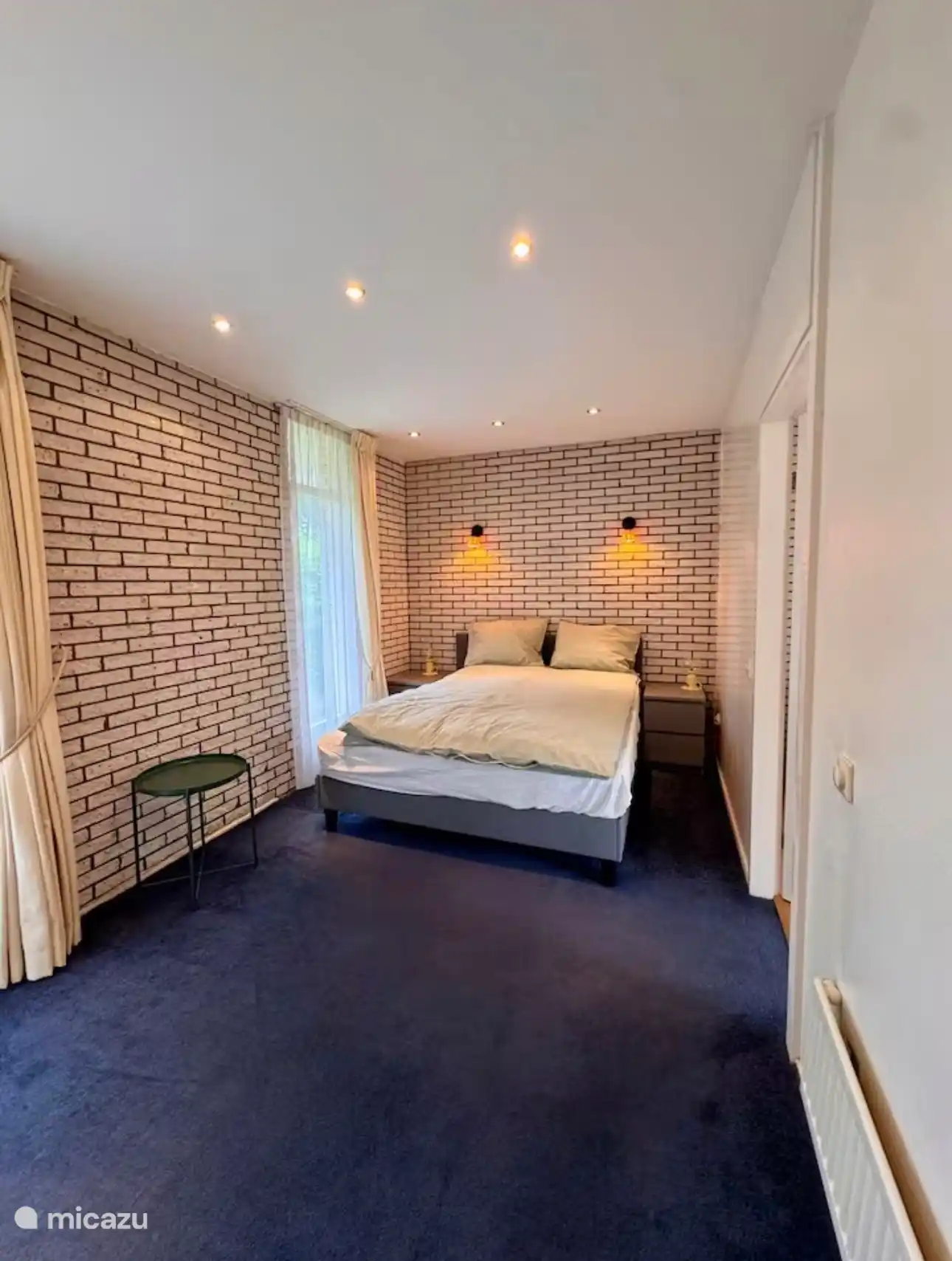 Rustige slaapkamer met comfortabele boxspring en sfeervolle verlichting. De lichte tinten en bakstenen muur geven de ruimte een warme en ontspannen uitstraling.