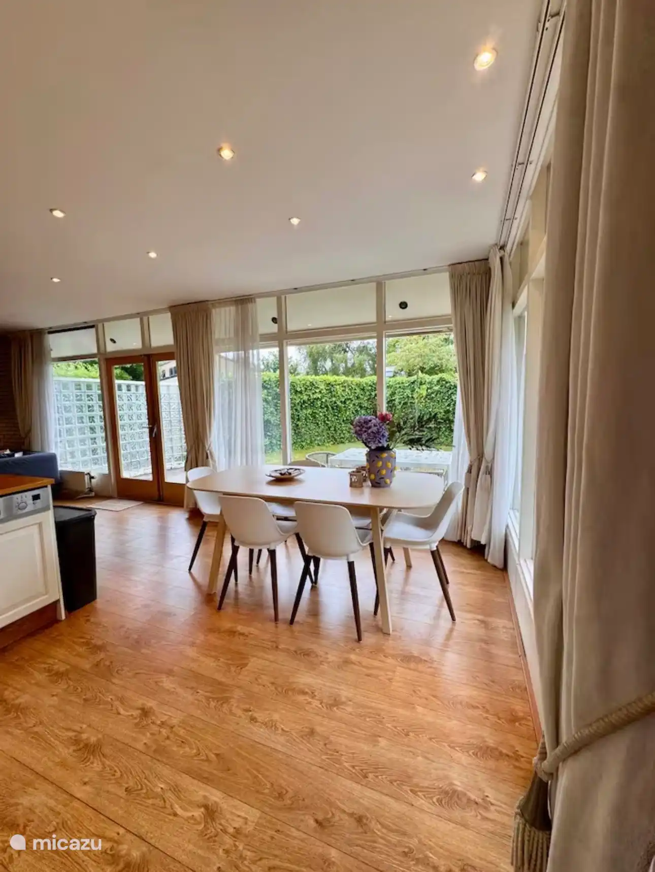 Lichte eetkamer met grote ramen en uitzicht op de groene tuin. Het zachte daglicht en de warme houtvloer creëren een open en uitnodigende plek om samen te genieten.