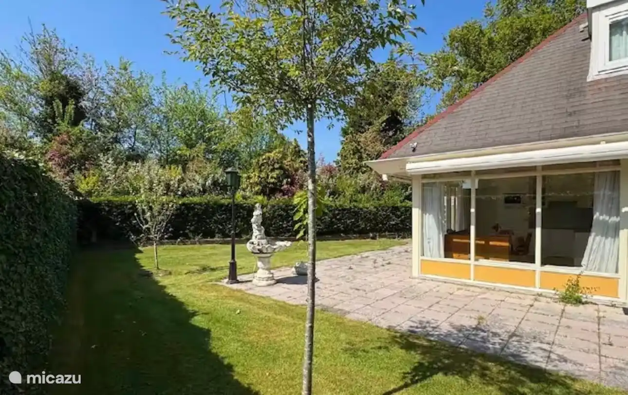 Ruime tuin met zonnig terras en groene beplanting. De rustige omgeving en het uitzicht op het charmante huis maken dit de perfecte plek om buiten te ontspannen.