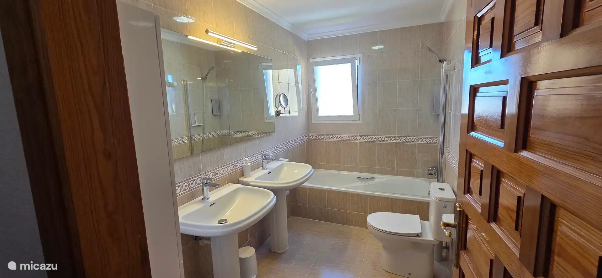 Ruime badkamer met twee wastafels, een ligbad/douche, toilet en kast. Haardroger, zeep, shampoo en douchegel dispensers aanwezig.