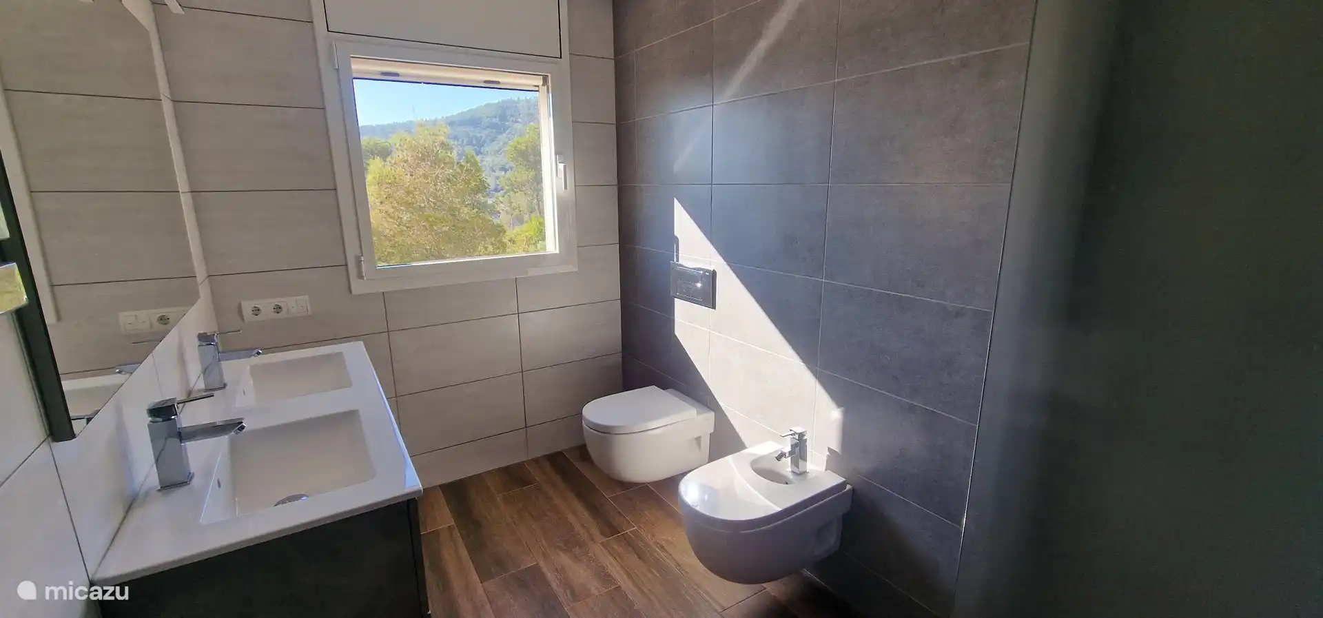 Grijze badkamer met toilet en bidet, douche, wastafelmeubel met handdoeken. Shampoo, conditioner, douchegel en toiletpapier aanwezig.