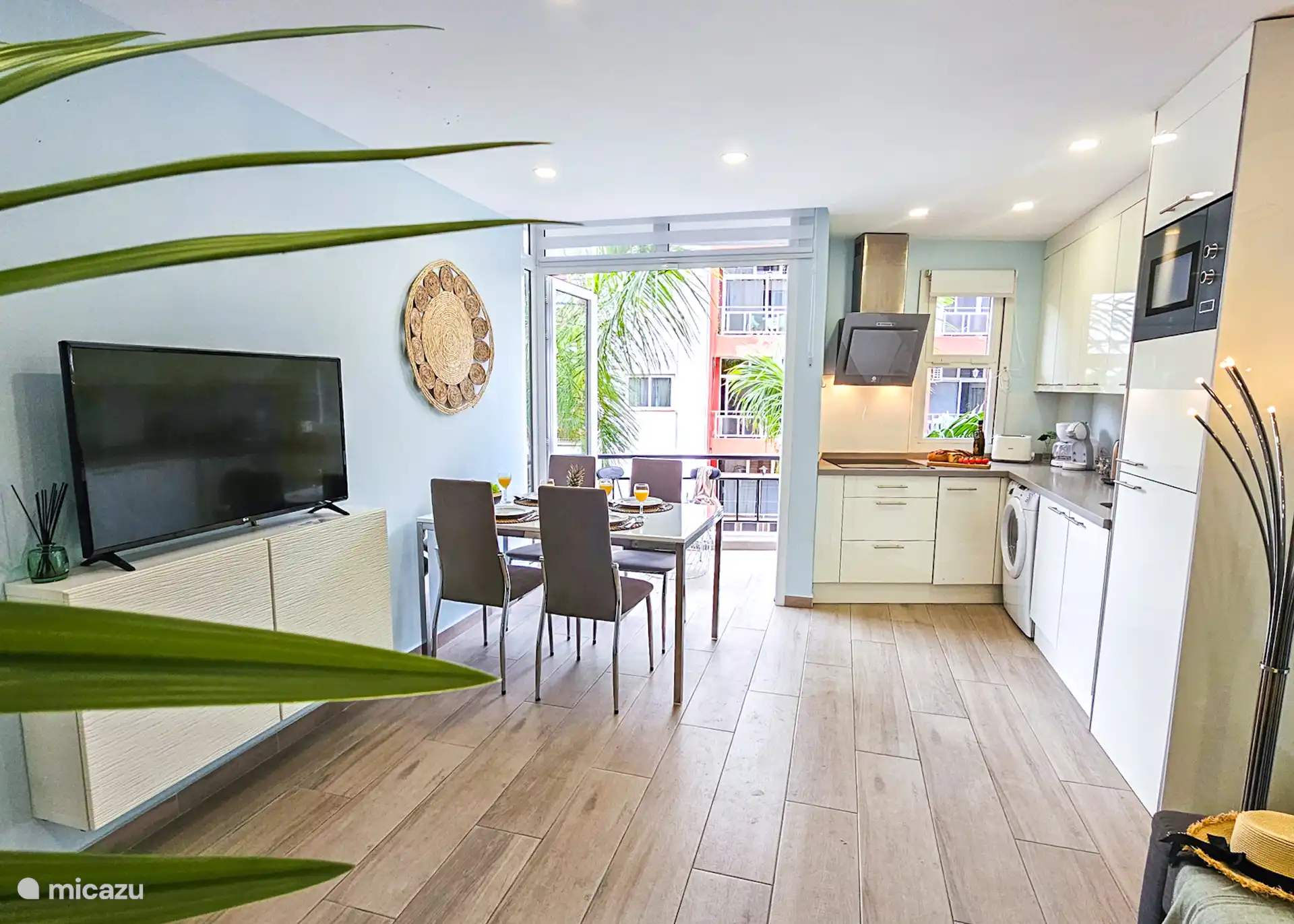 Gezellige open woonkamer en keuken met veel licht. Ramen bieden uitzicht op palmbomen langs de weg naar het strand. Geniet van maaltijden aan de eettafel, met moderne gemakken binnen handbereik.