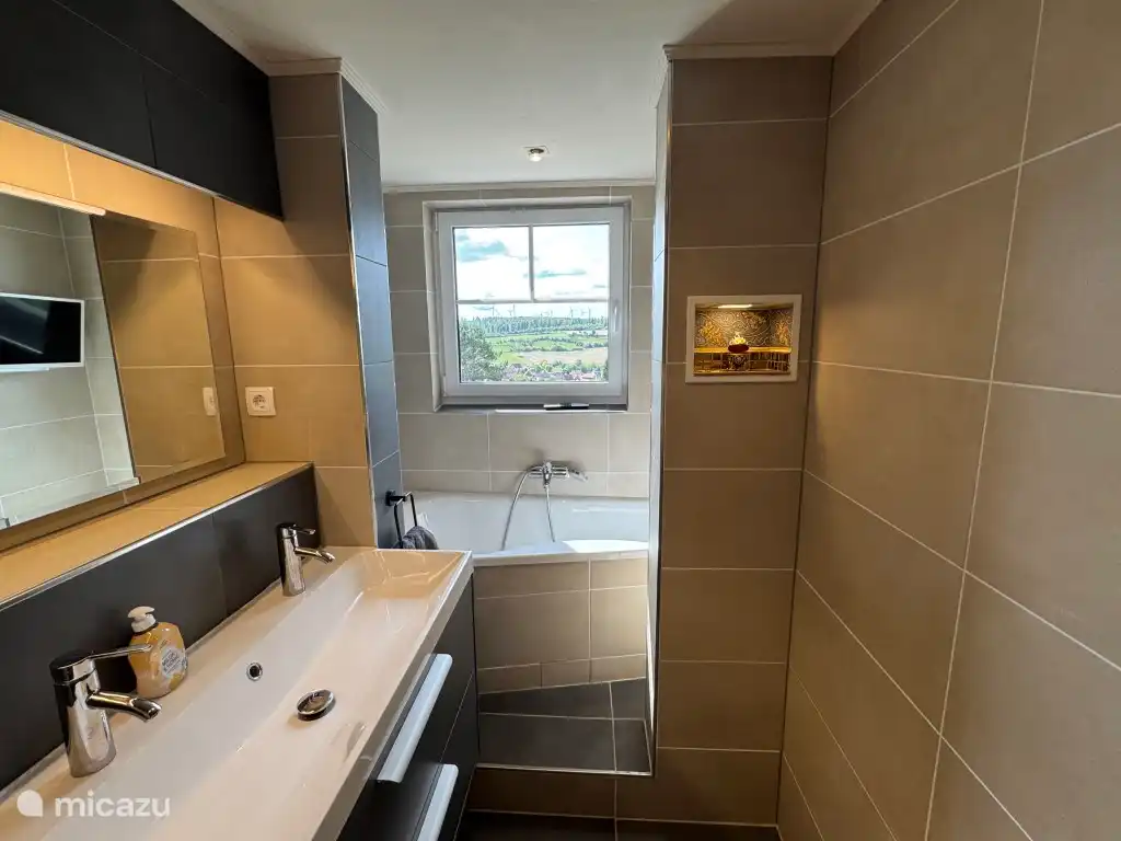 De grote badkamer met ingebouwd bad, toilet en dubbele wastafel