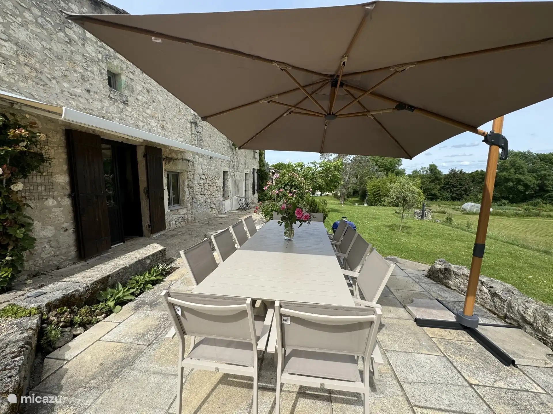 Terraza en la parte delantera de la casa con mesa de comedor, parasol, barbacoa y vistas al jardín y al campo circundante.