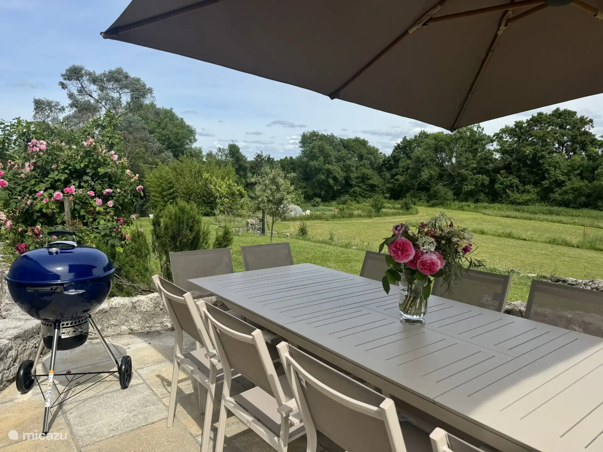 Terraza en la parte delantera de la casa con mesa de comedor, parasol, barbacoa y vistas al jardín y al campo circundante.