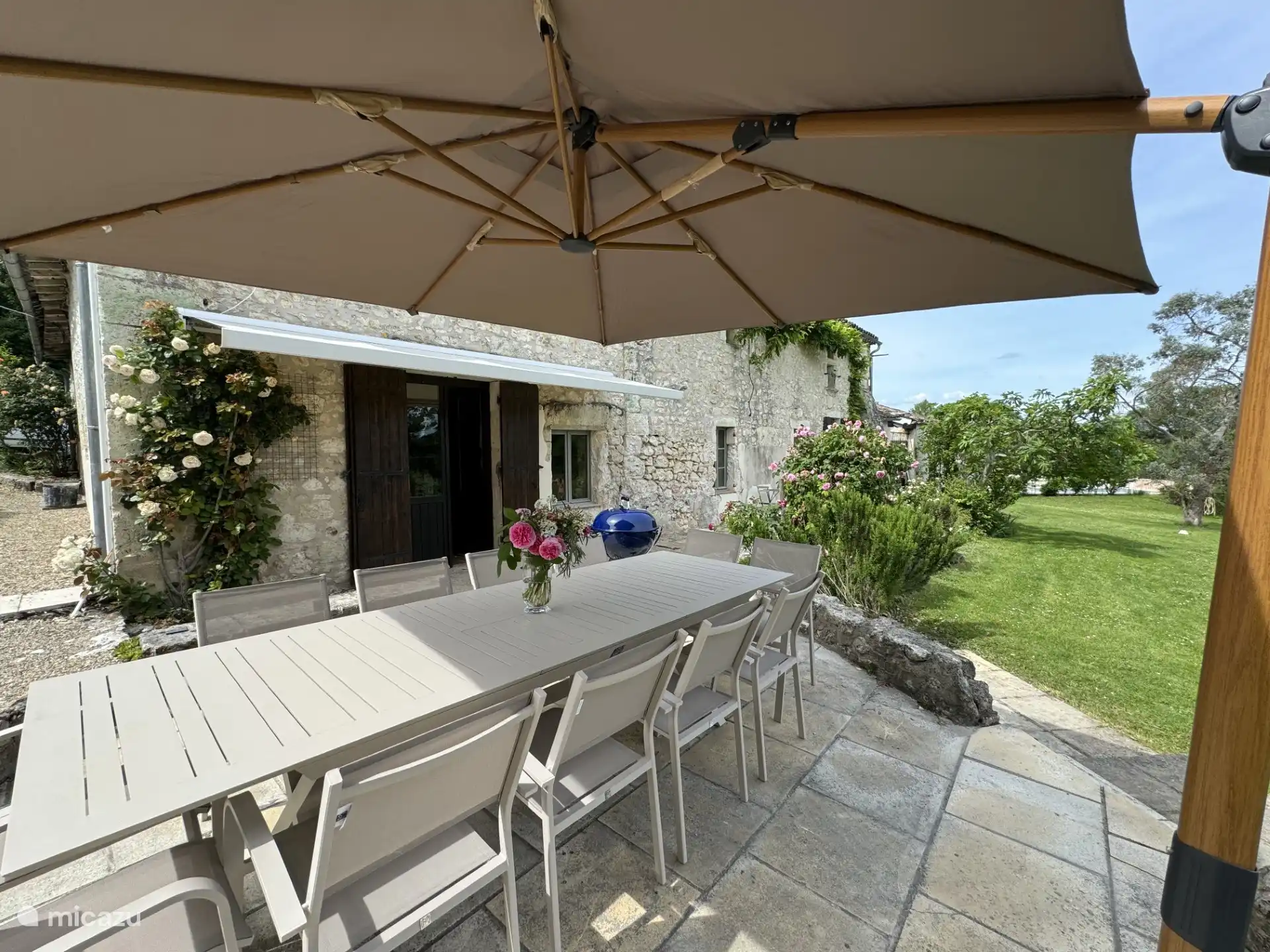 Terraza en la parte delantera de la casa con mesa de comedor, parasol, barbacoa y vistas al jardín y al campo circundante.