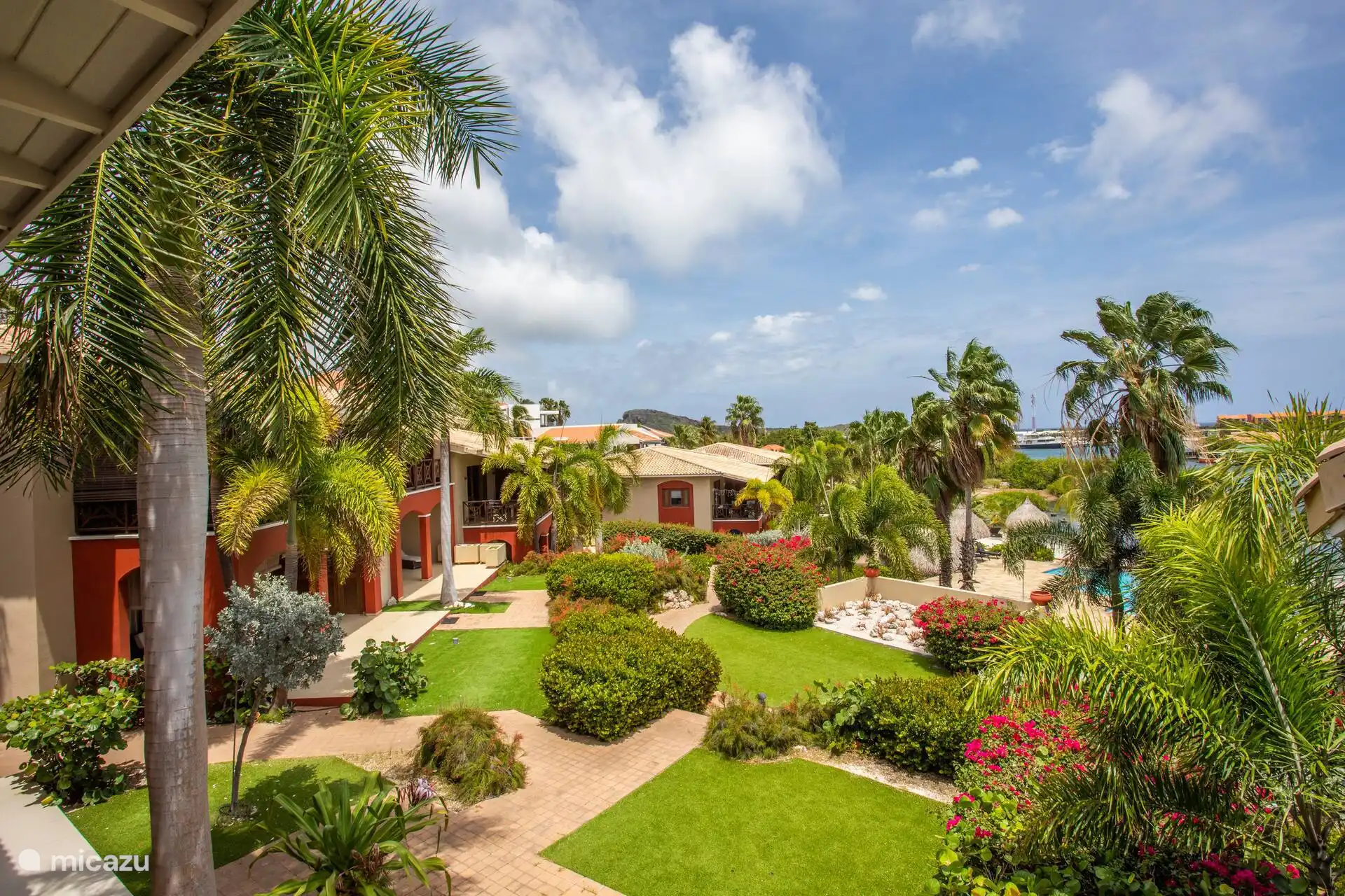 Blick über den tropischen Garten des Resorts mit Palmen, bunten Pflanzen und dem Schwimmbad.