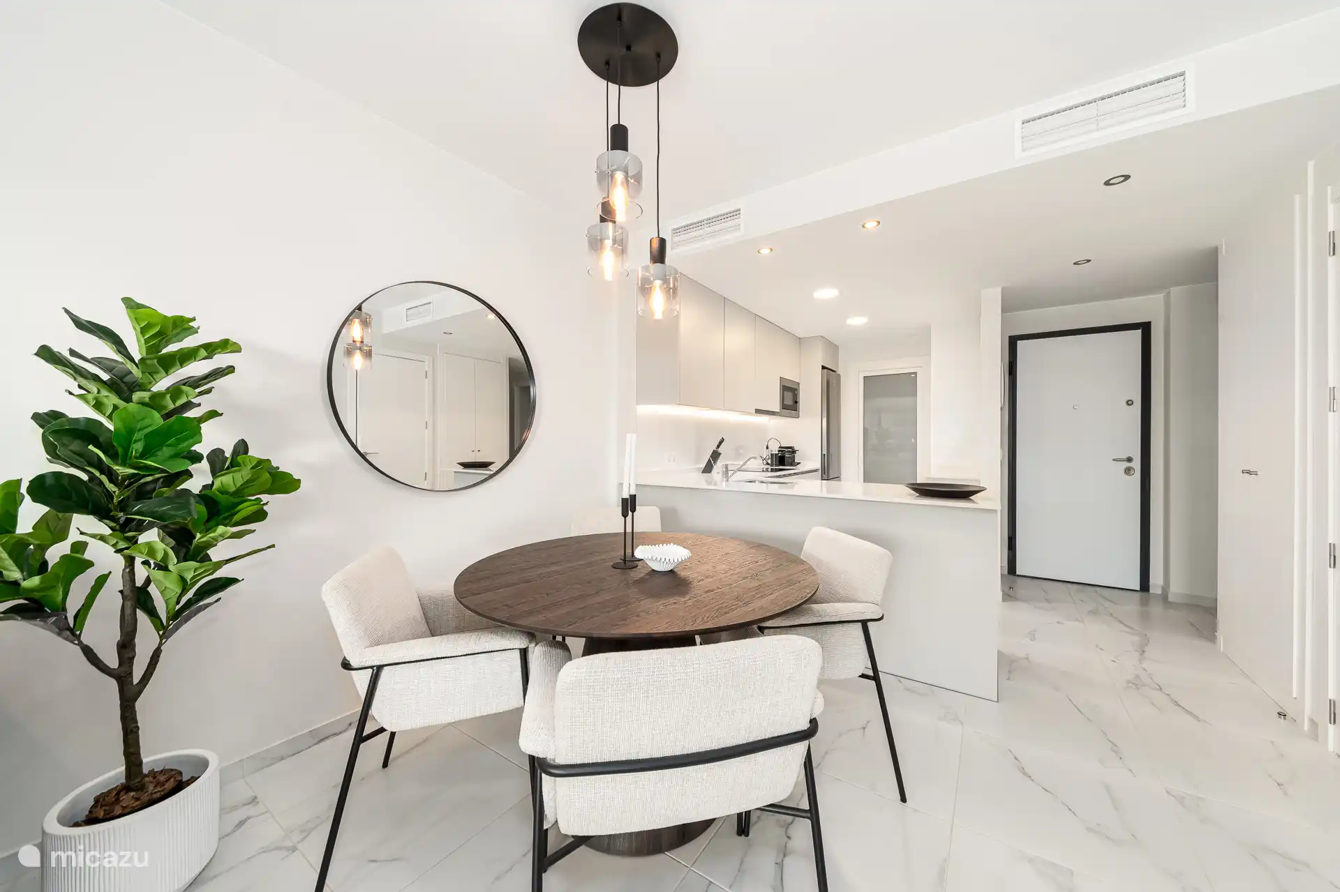 Eetkamer met ronde houten tafel, stoffen stoelen en hanglamp, met open keuken op de achtergrond.
