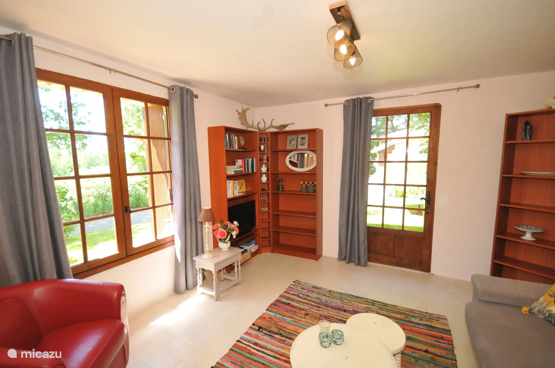 Salon avec smart TV, deux fauteuils, canapé-lit et porte donnant sur le jardin et la candaquine.