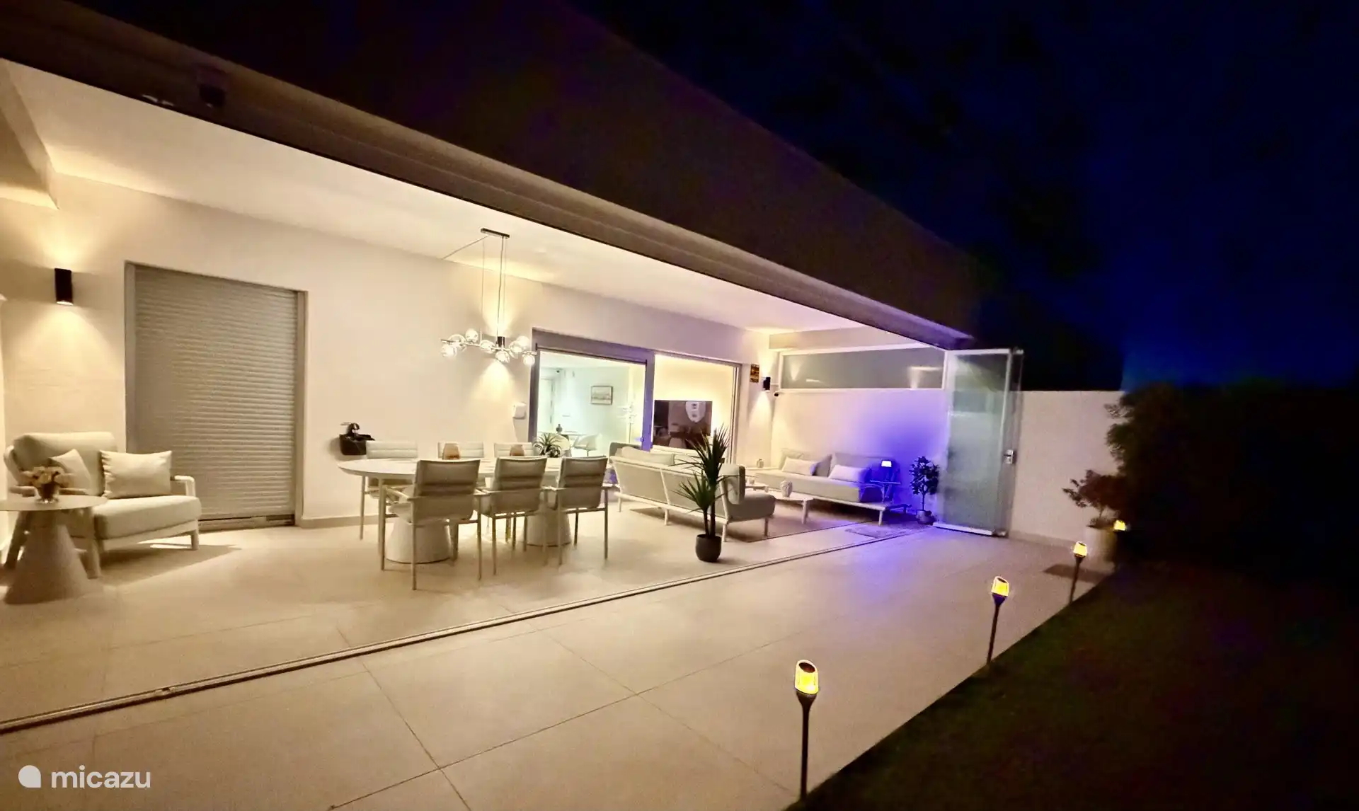 The terrace in the evening with the lights on: the spacious dining table, the lounge sofa and the passage to the living room.