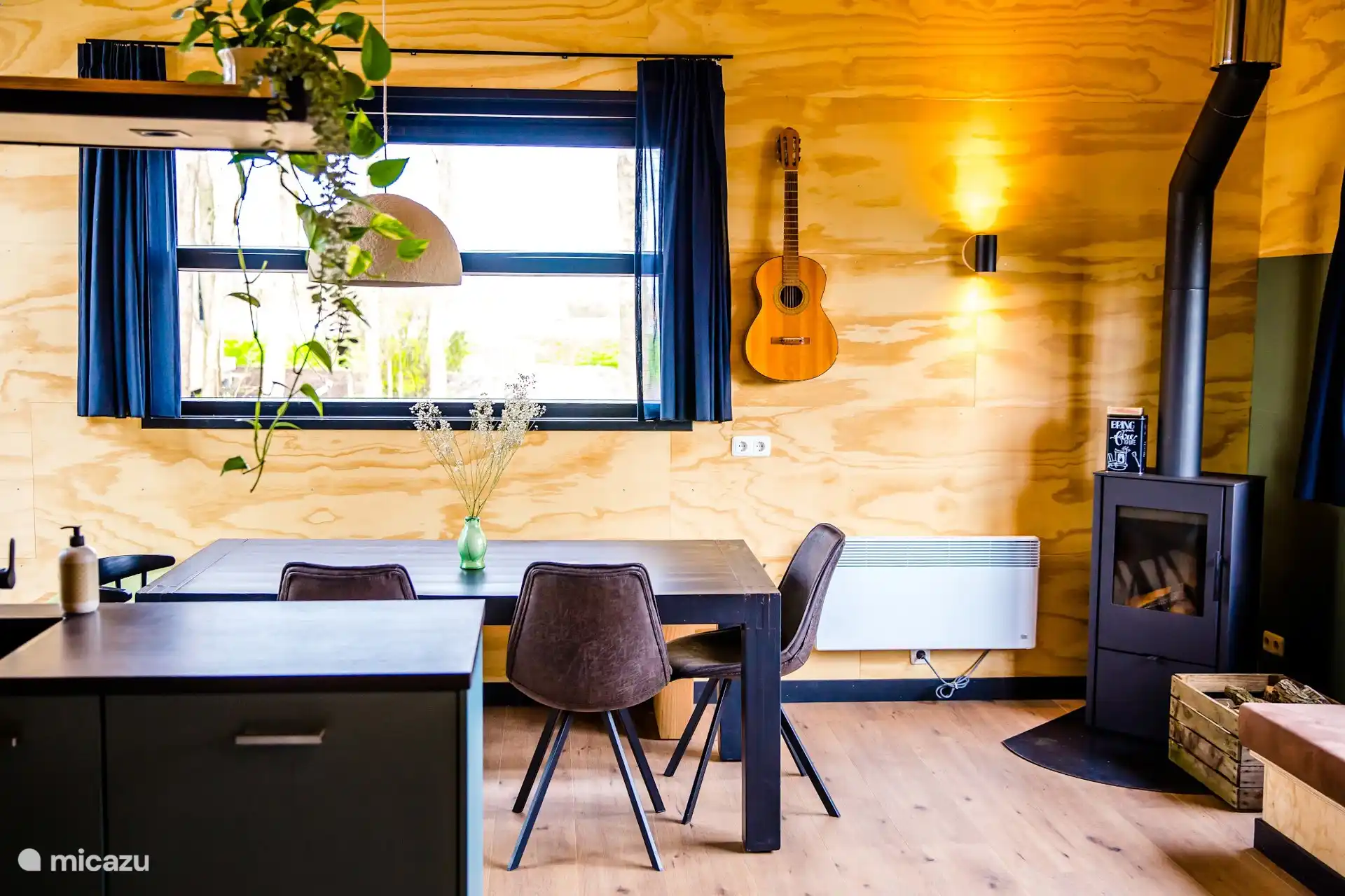 Open woonkamer met houtkachel, eettafel en gitaar aan de wand voor een sfeervolle zithoek.