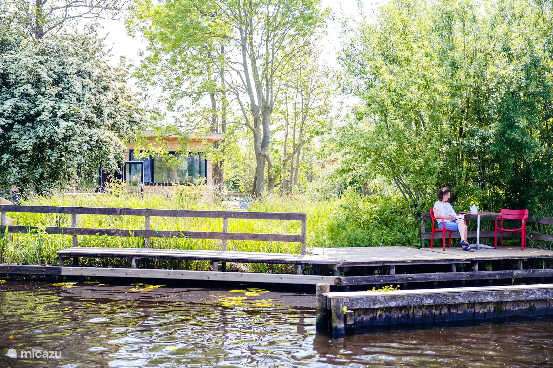 Houten vlonder aan het water bij Waterlodge Iisfûgel, met zicht op het terras van de lodge tussen het groen.