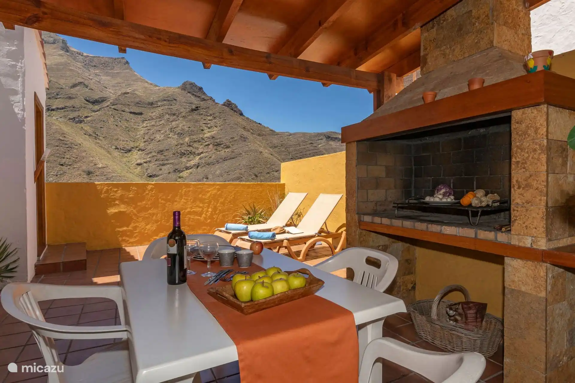Terraza cubierta con barbacoa de obra, mesa y tumbonas al fondo.