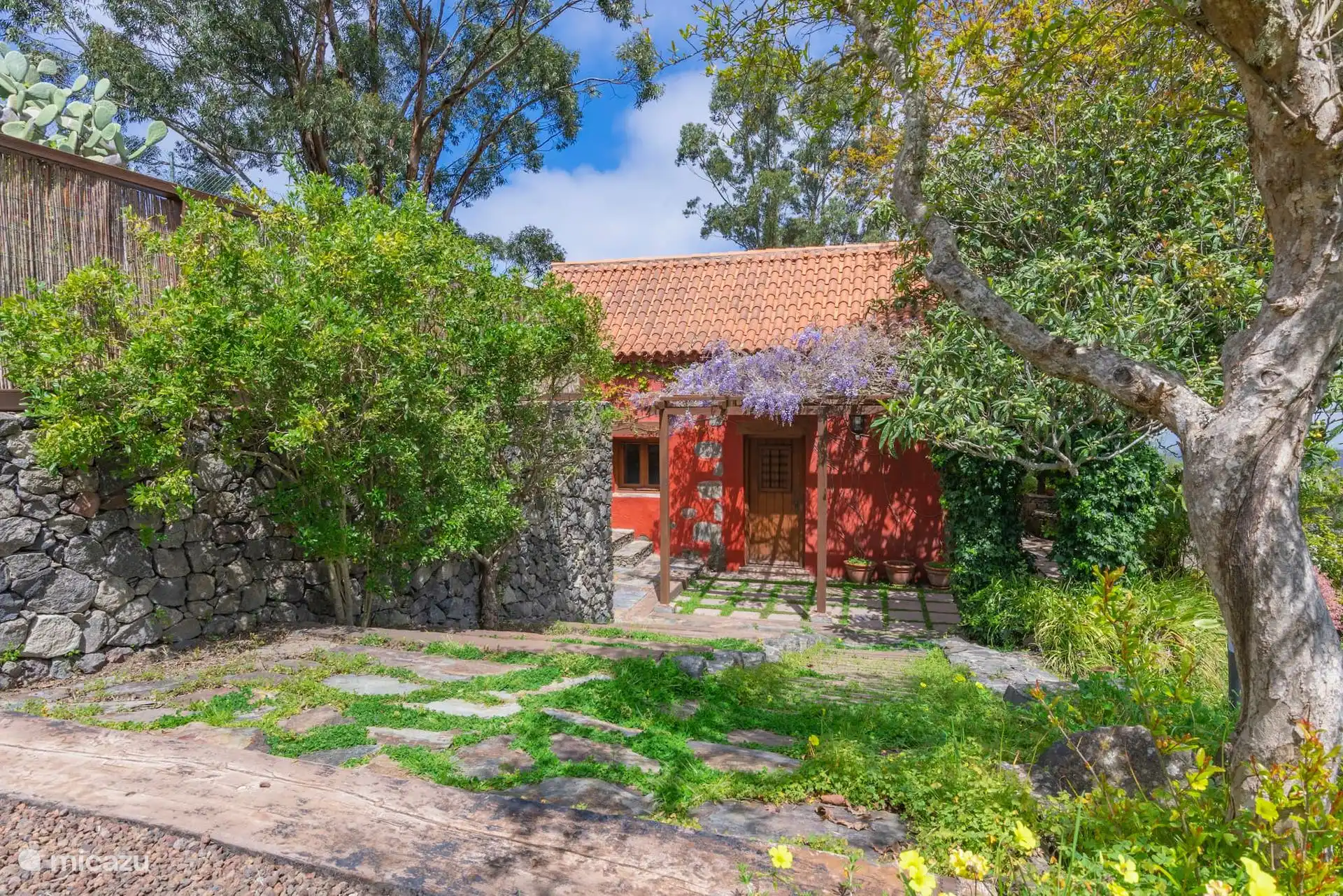 El jardín con muros de piedra y la casa roja entre los árboles.