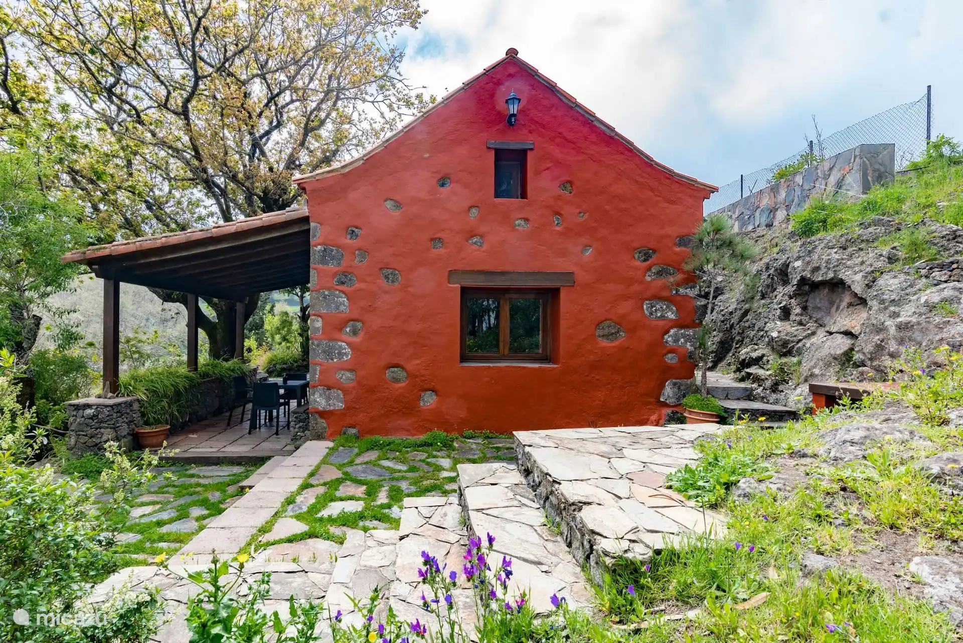 La casa centenaria de piedra roja, con el porche y el jardín delante.
