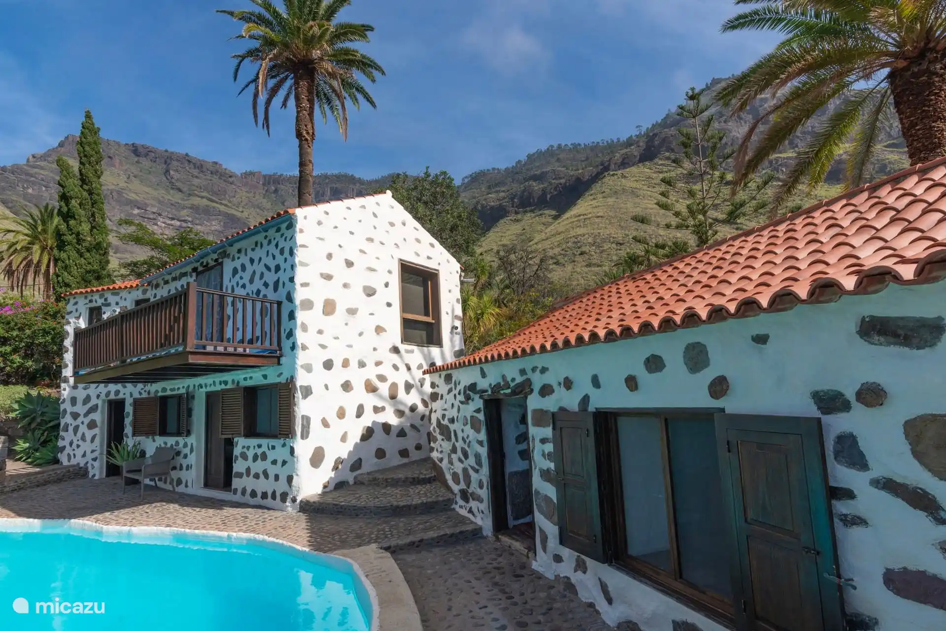 Fachada de la casa con la piscina delante y el risco detrás.