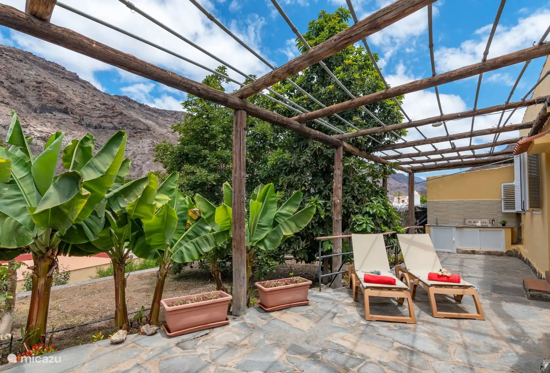 Eine Terrasse unter der hölzernen Pergola mit Liegeplätzen, Bananenbäumen und der Außenküche der Finca.
