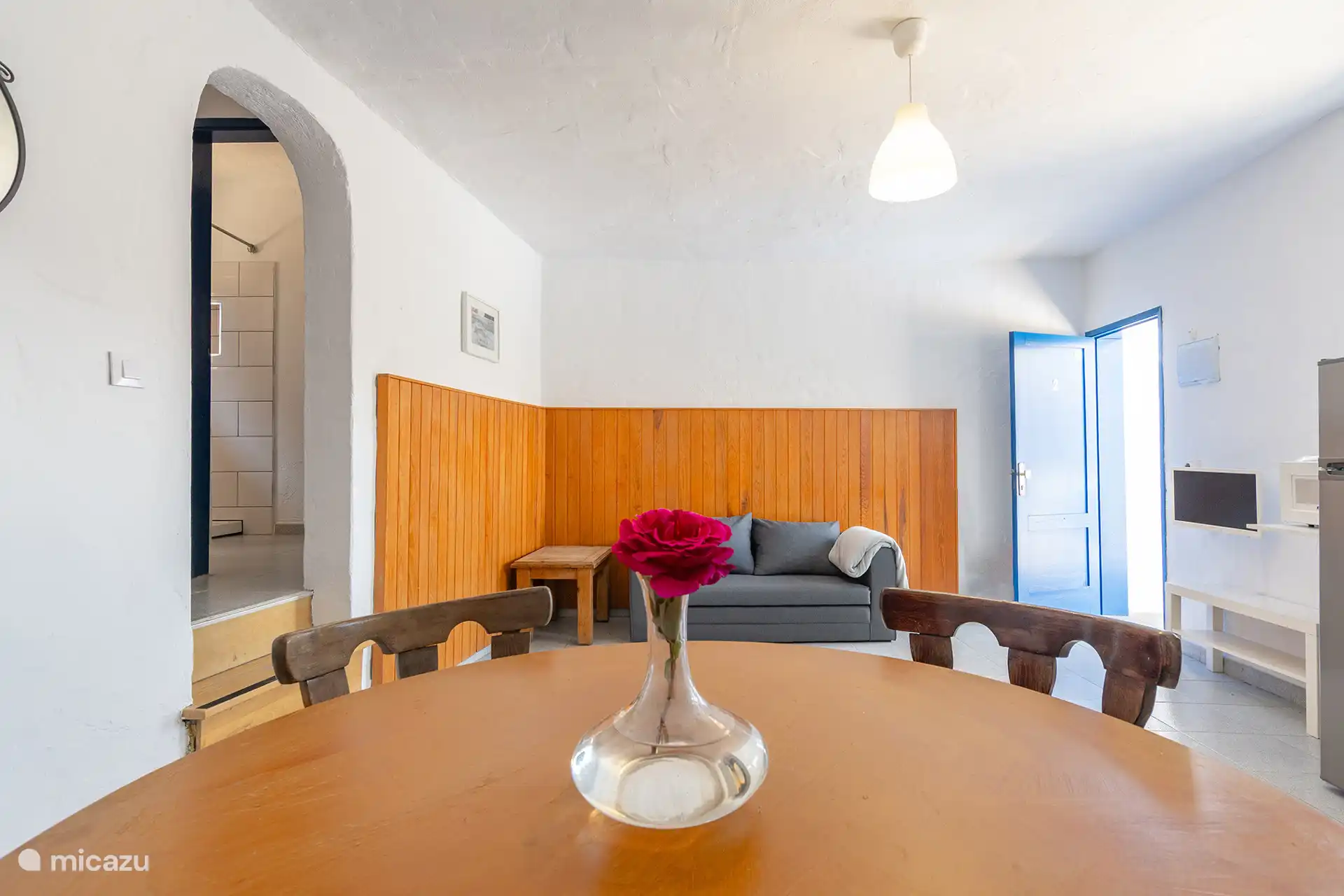 Woonkamer met slaapbank en tafel voor vier personen, met de doorgang naar de badkamer aan de linkerkant.
