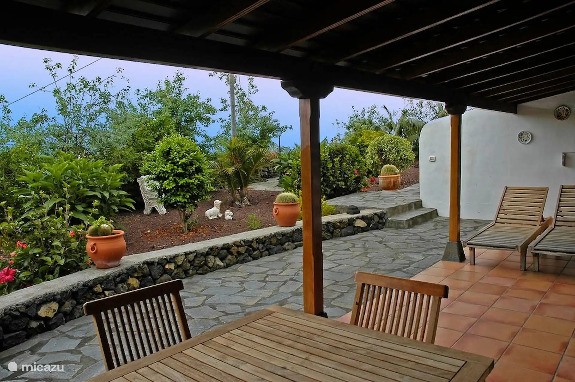 Veranda met tafel, stoelen en ligstoelen, open op de cactustuin en de vulkanische stenen muur.