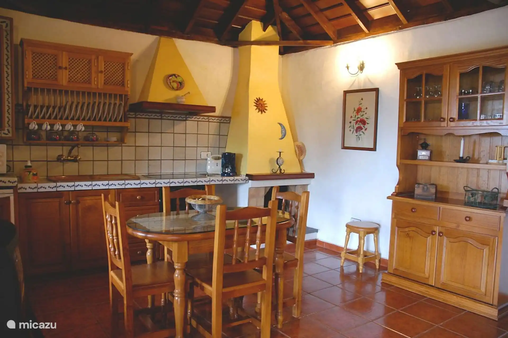 Keuken en eetkamer onder het houten dak, met open haard, kast en servies.