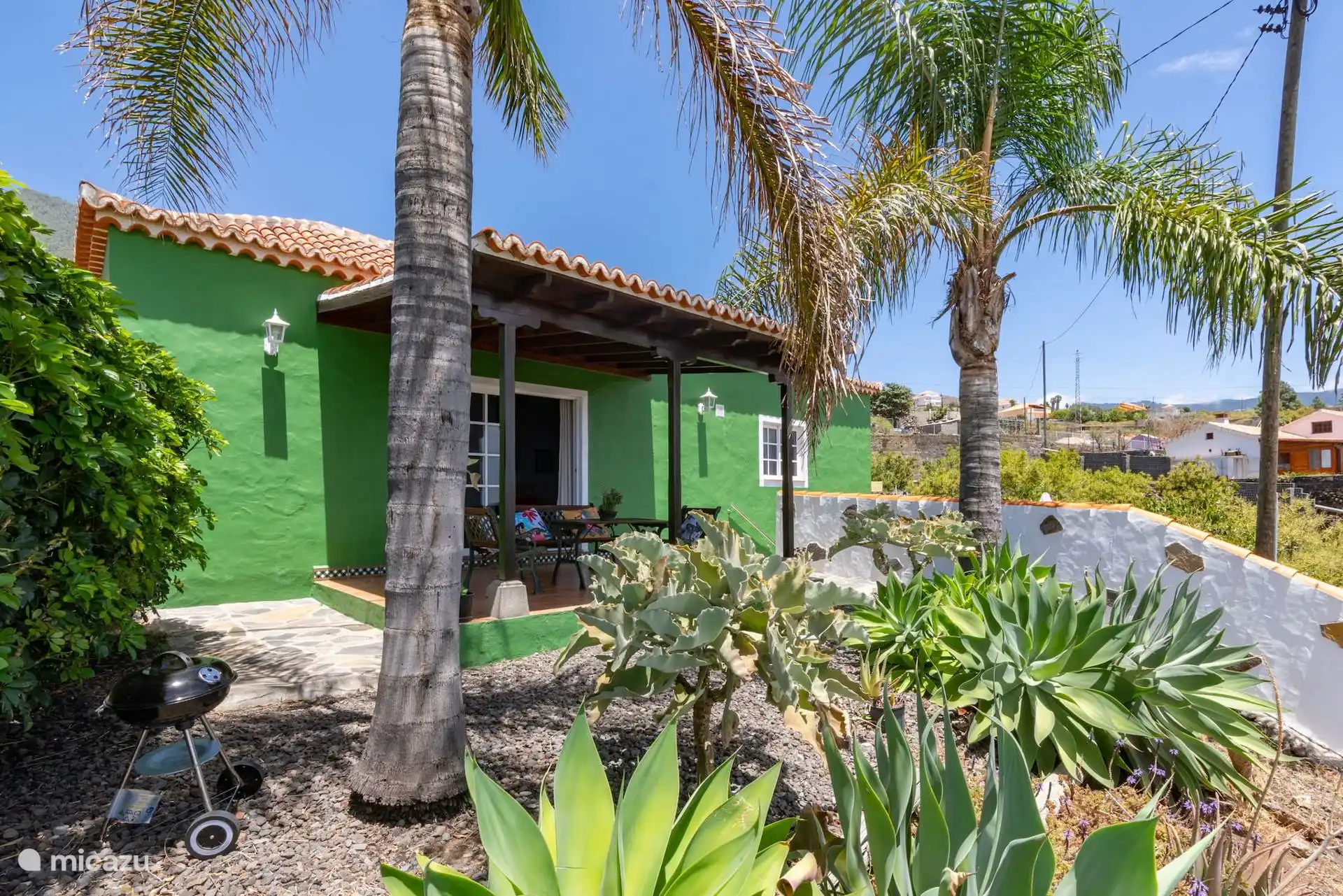 Groene gevel met overdekte veranda en tafel; Voor de deur de grindtuin met palmbomen, agaven en een draagbare barbecue.