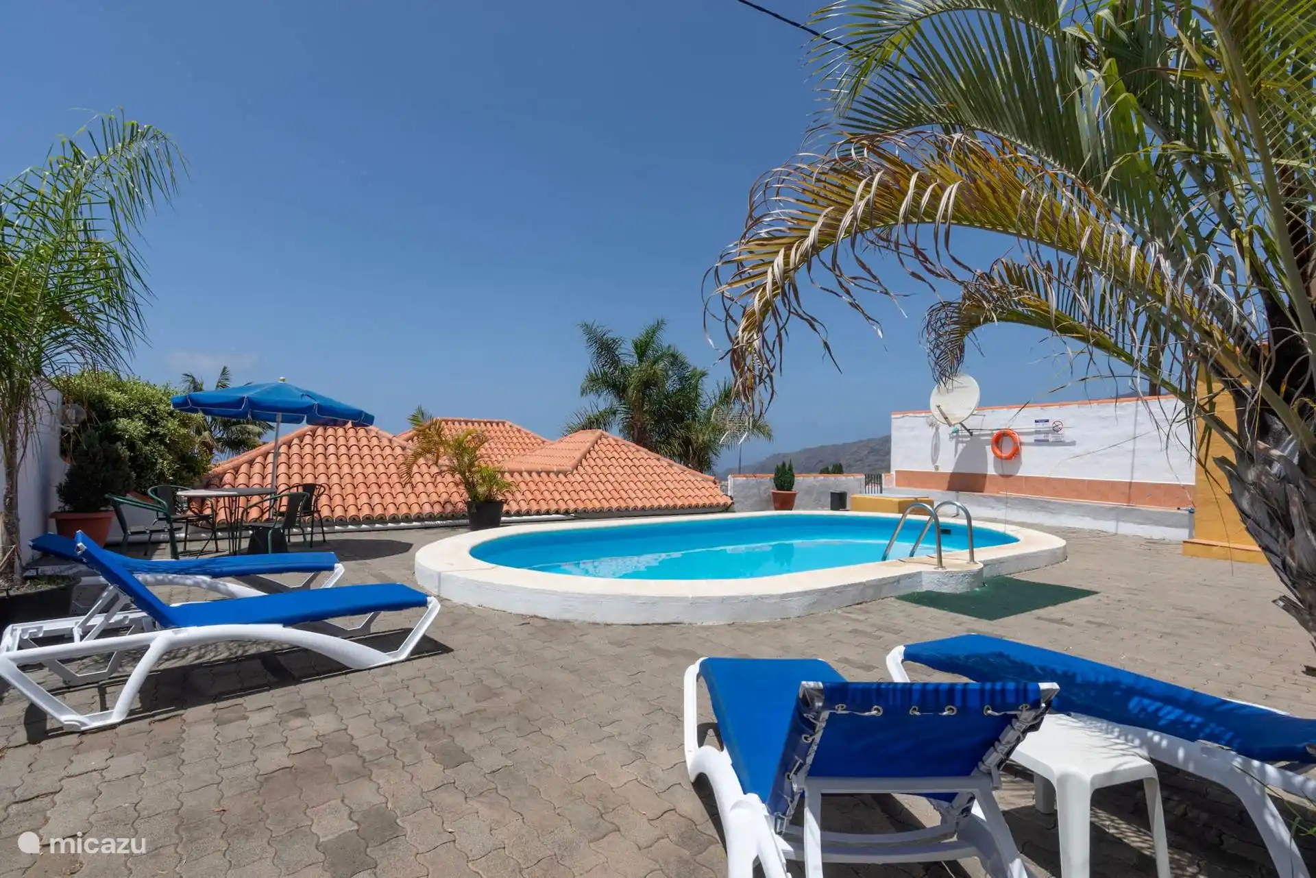 La piscine et le solarium, avec des chaises longues et une table avec des chaises contre le mur ; derrière, les toits et la montagne.