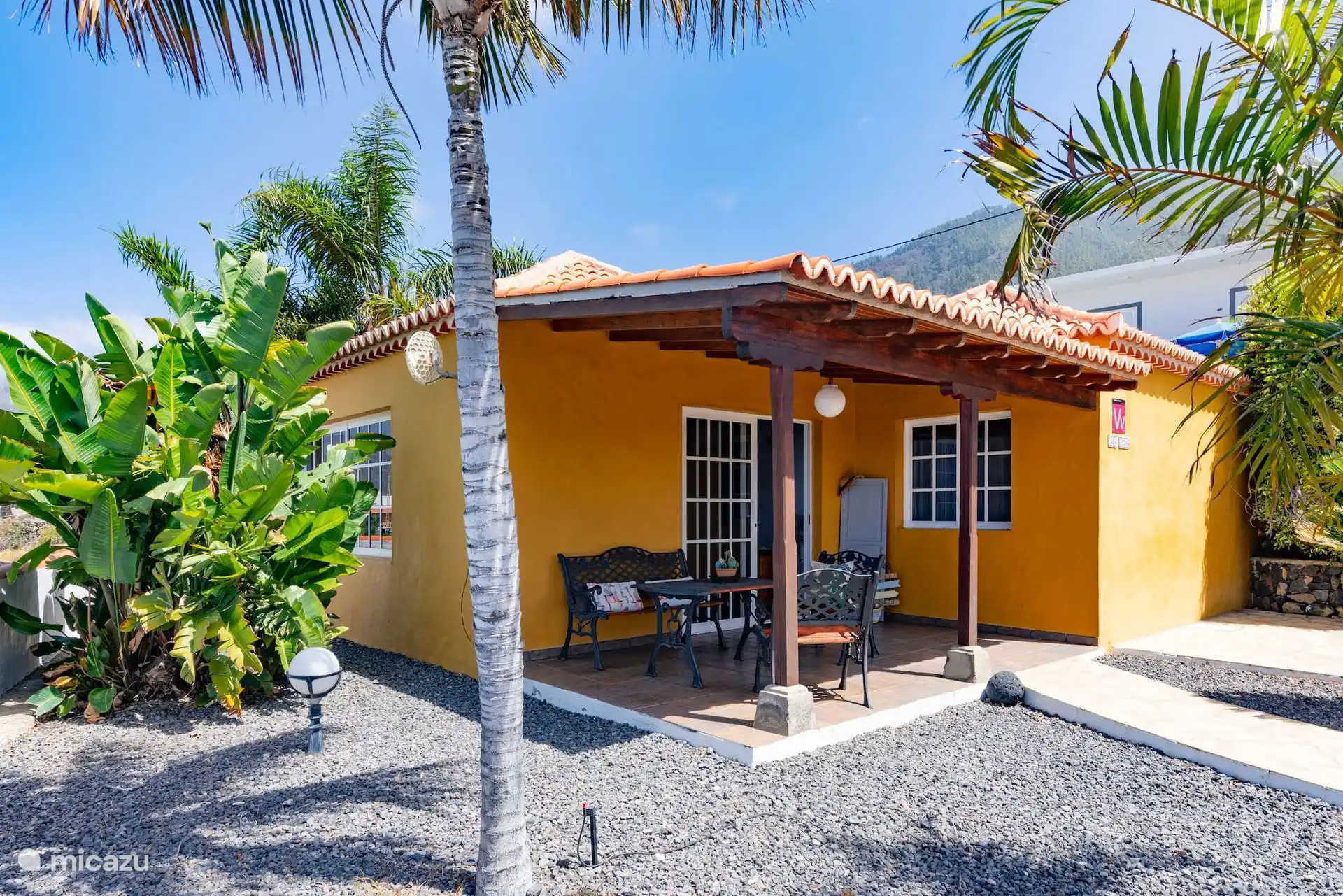 La maison à l’extérieur : façade jaune, toit en tuiles et porche d’entrée avec table et bancs en fer forgé, parmi les palmiers.