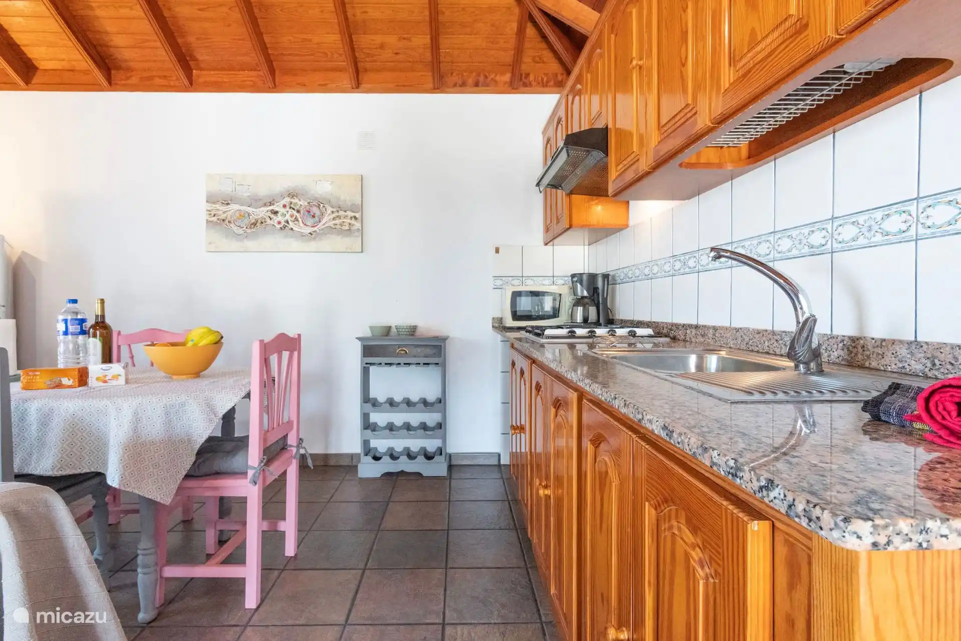 Cuisine en bois avec un plan de travail en granit, un évier, une cuisinière à gaz, un micro-ondes et une cafetière ; à côté, la table à manger et le porte-bouteilles.