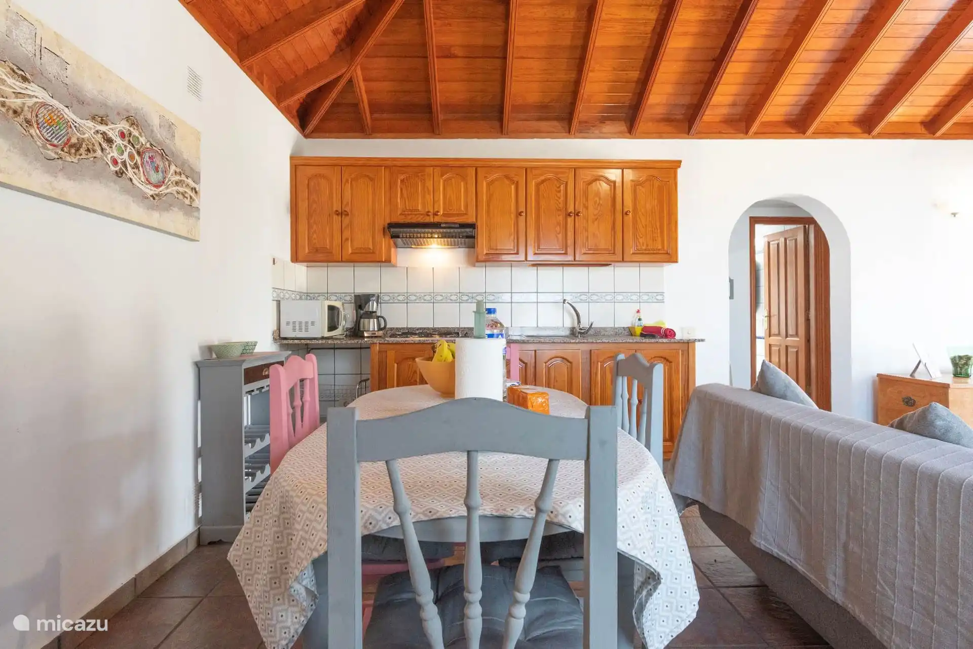 Salle à manger et cuisine dans la même pièce : table avec chaises, meubles en bois, micro-ondes et hotte ; à droite, l’arche du couloir.