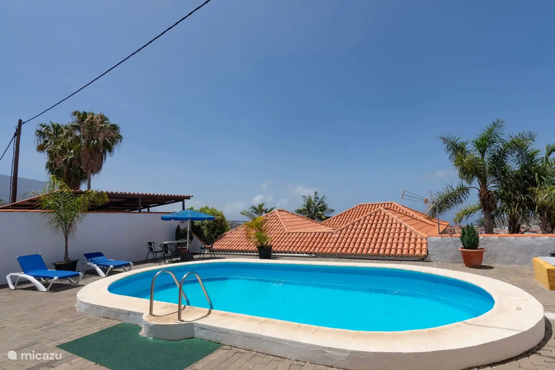 La piscine avant, avec l’échelle d’accès, les transats et le parasol ; derrière, les toits et les palmiers.