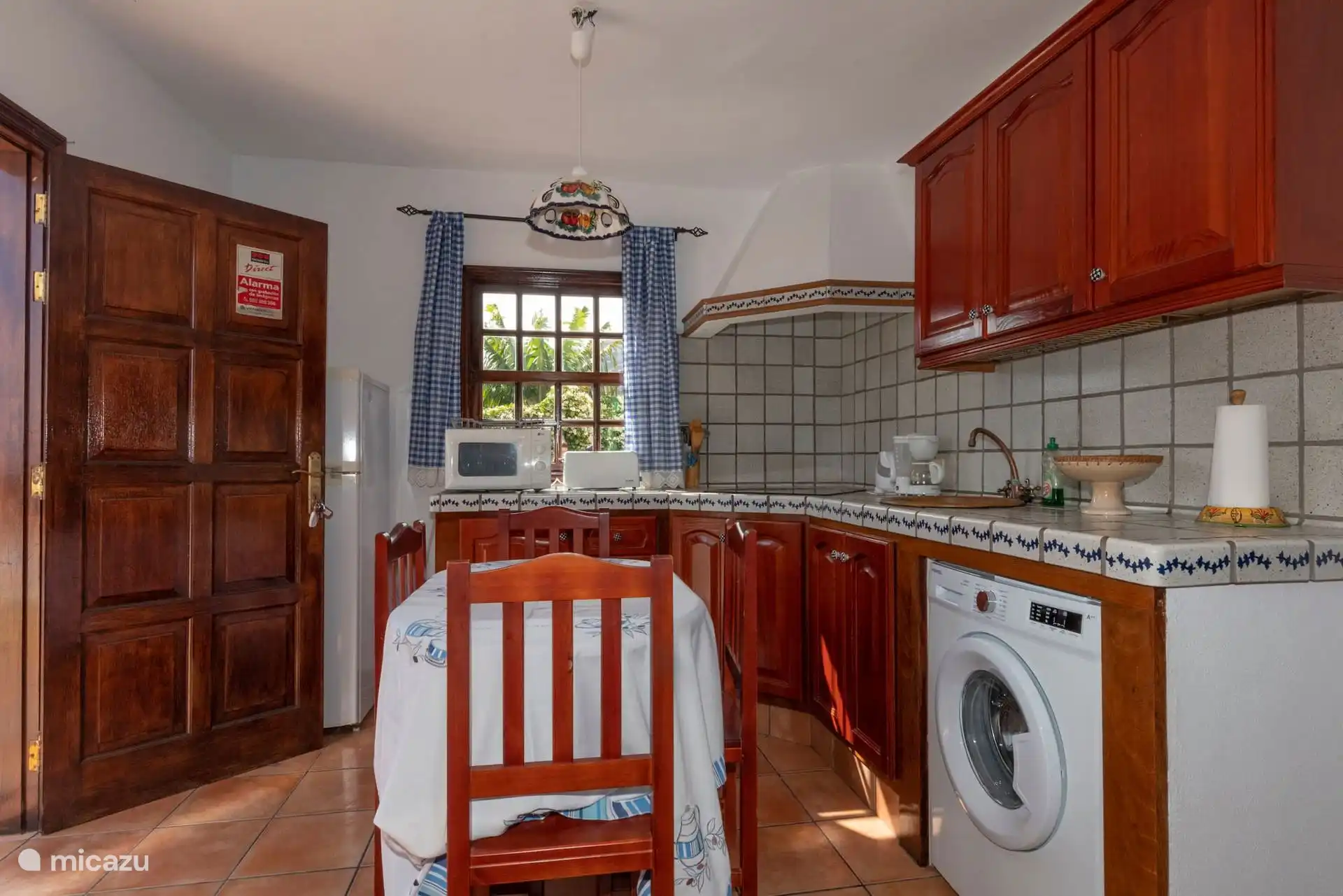 Kitchen with wooden furniture and tiled worktop: microwave, toaster, coffee maker, fridge and washing machine; Table with chairs by the window.