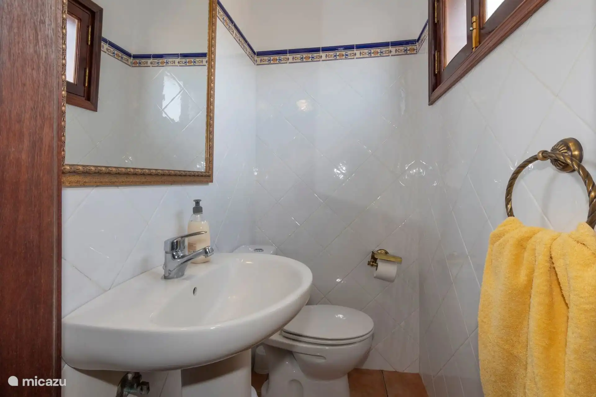 The toilet of the two-bed bedroom, with sink, toilet and mirror, tiled in white with blue border.