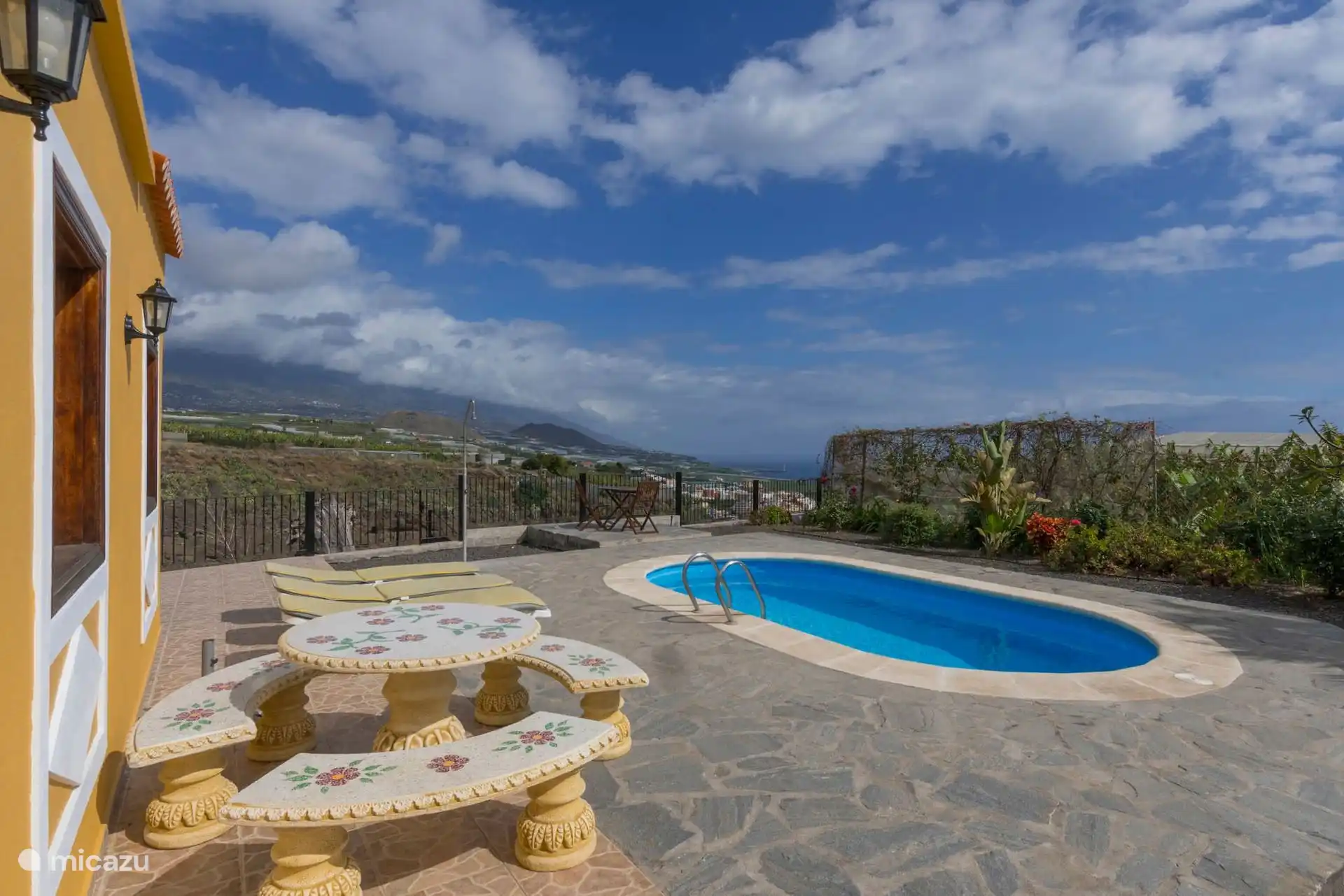 Stone table with poolside benches; beyond the railing, banana plantations, the town and the sea.
