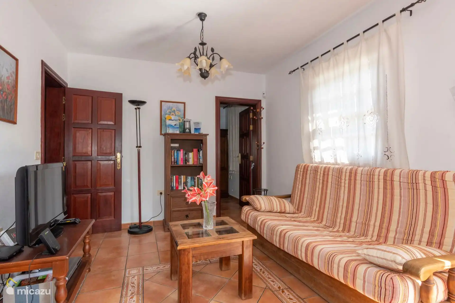 Living room with sofa, wooden coffee table and television; Bookshelf with books and clay floor.