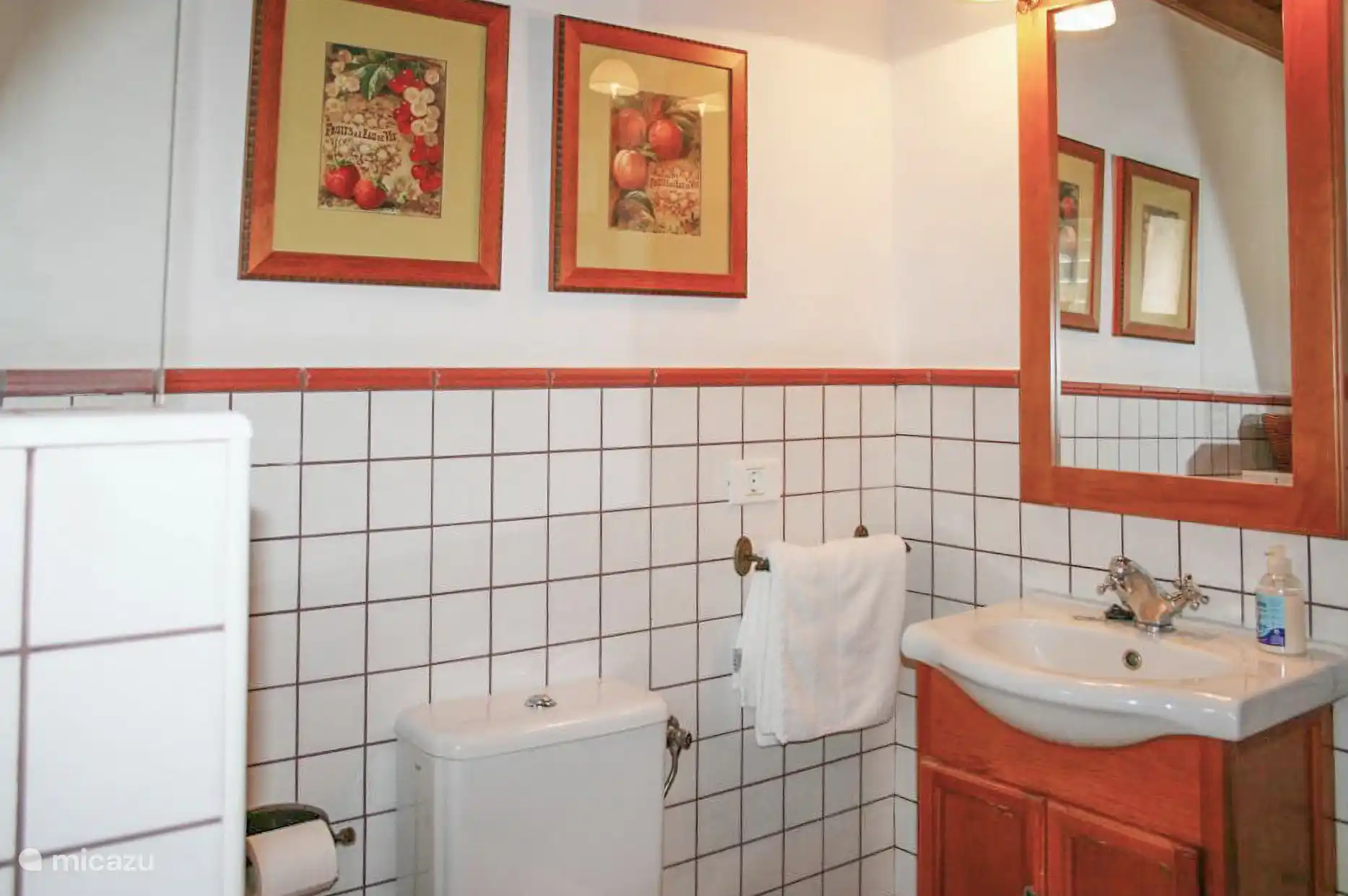 Witte betegelde badkamer, met wastafel op houten meubels, spiegel en toilet.