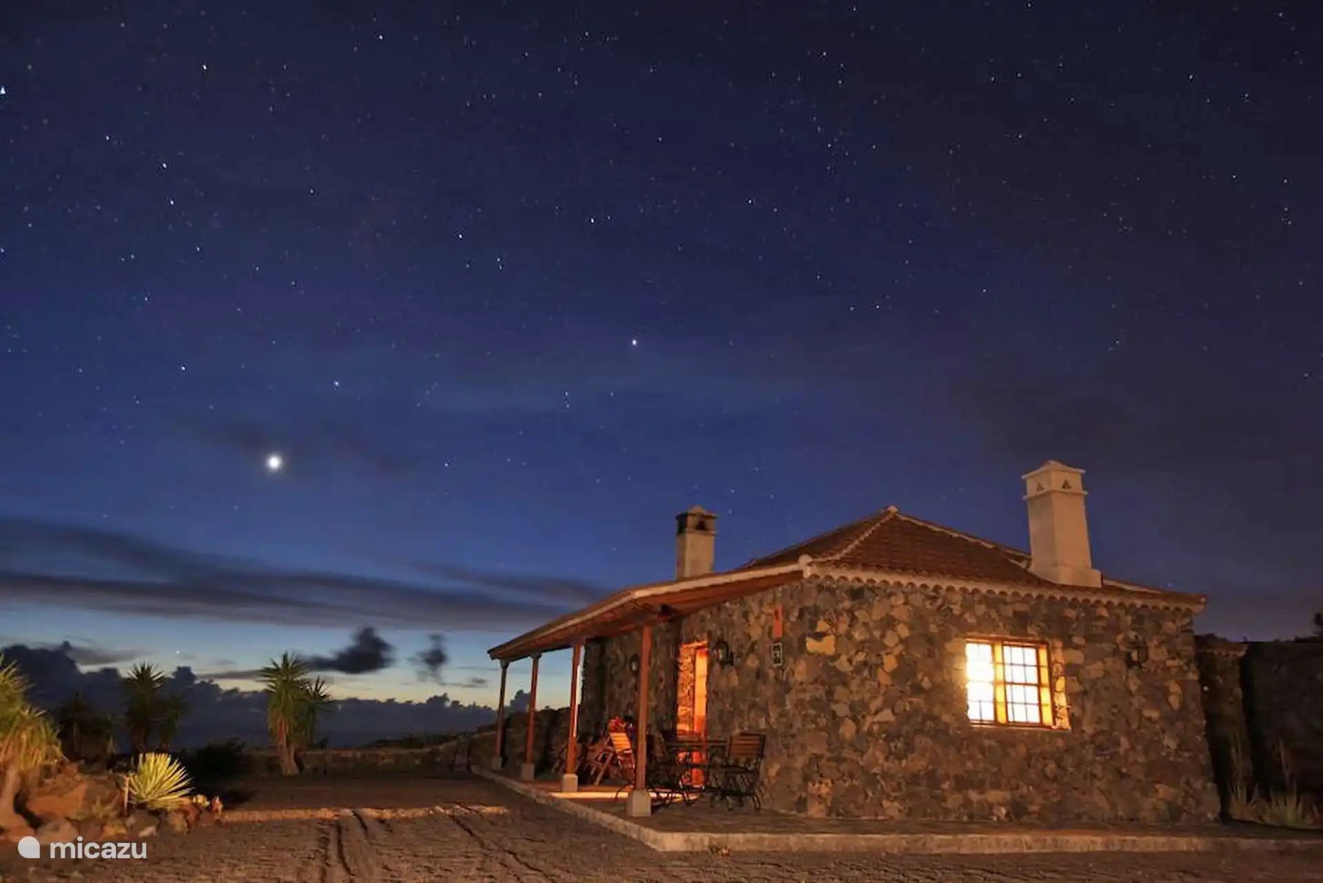 The house at night, with the light on in the window and the starry sky above it.
